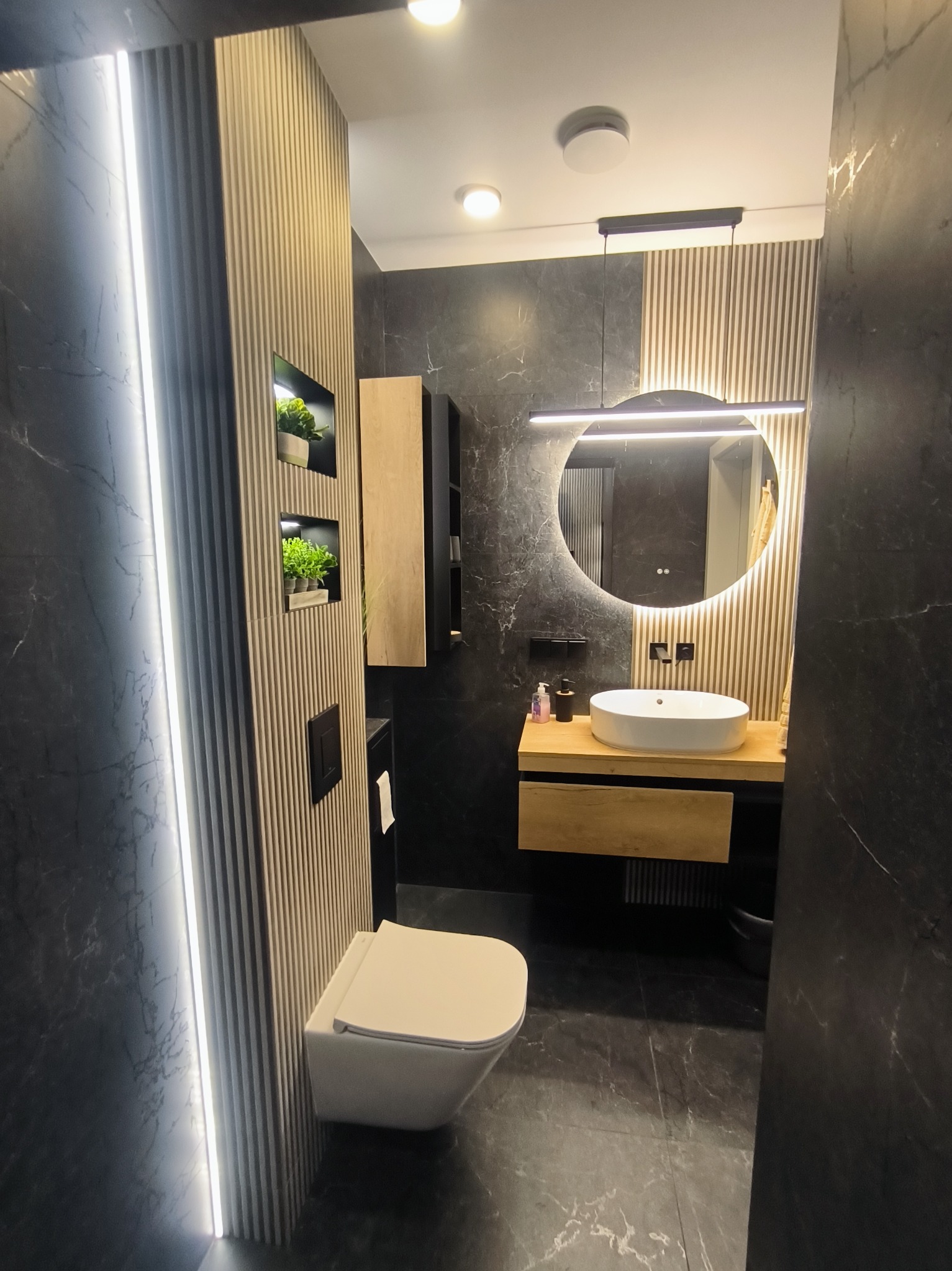 Nowoczesna łazienka w Wołominie z czarnymi płytkami imitującymi marmur, podświetlanym lustrem i drewnianymi akcentami. Minimalistyczny design z roślinami w niszach.