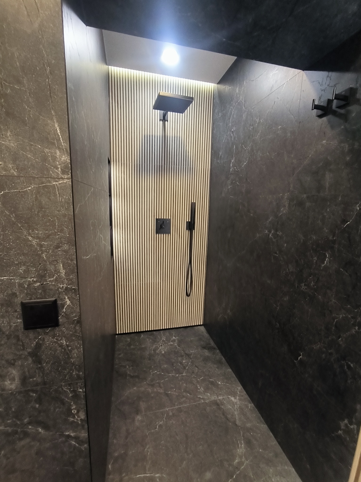 Nowoczesna kabina prysznicowa z ciemnymi płytkami imitującymi marmur i ścianą z drewnianych listew. Minimalistyczny design, czarna armatura.