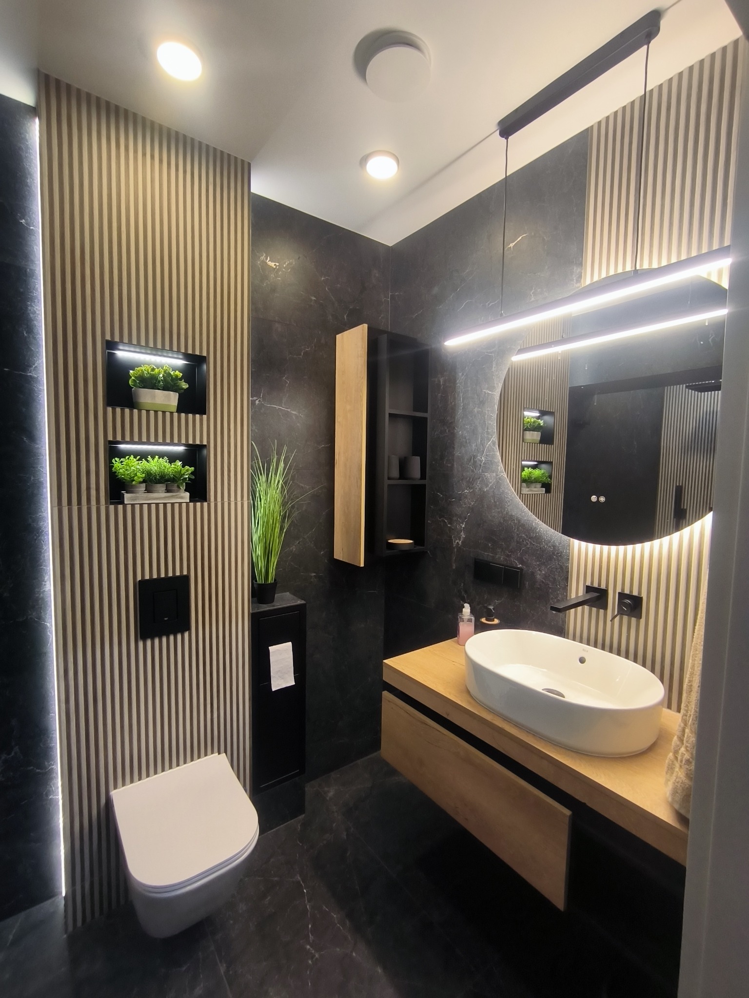 Nowoczesna łazienka z czarnymi marmurowymi ścianami, drewnianymi akcentami i okrągłym lustrem z podświetleniem LED. Uwagę przykuwają rośliny w niszach ściennych.