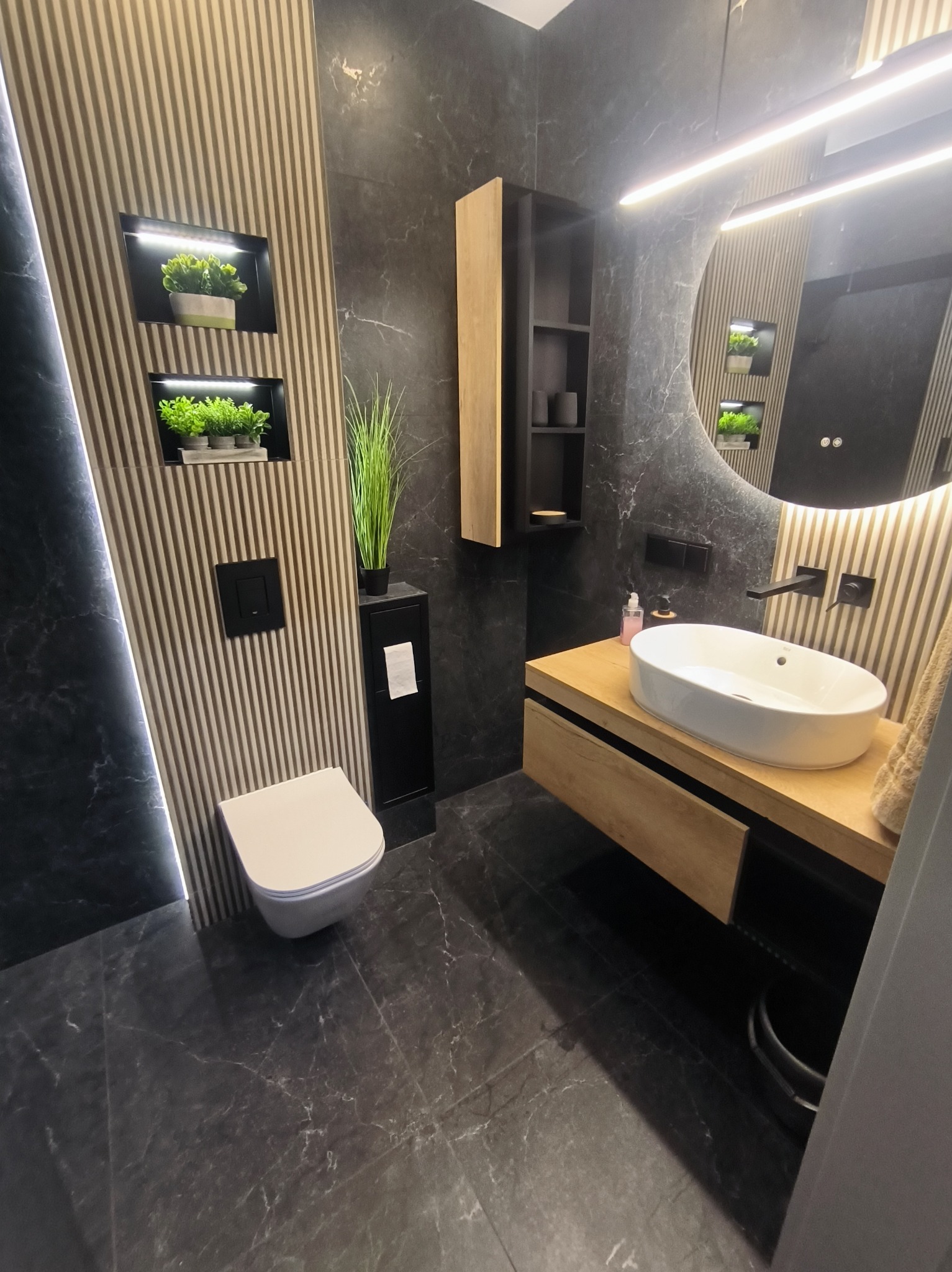 Nowoczesna łazienka z czarnymi płytkami imitującymi marmur, drewnianą szafką pod umywalką i lamelami na ścianie. Uwagę przykuwają rośliny wbudowane w ścianę.