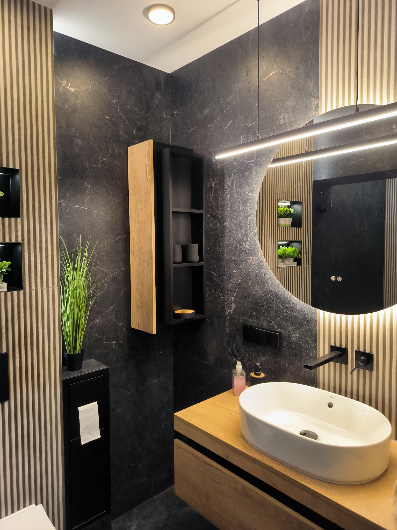 Nowoczesna łazienka z czarnymi marmurowymi płytkami, drewnianymi akcentami i okrągłym lustrem z podświetleniem LED. Minimalistyczny design, eleganckie wykończenie.