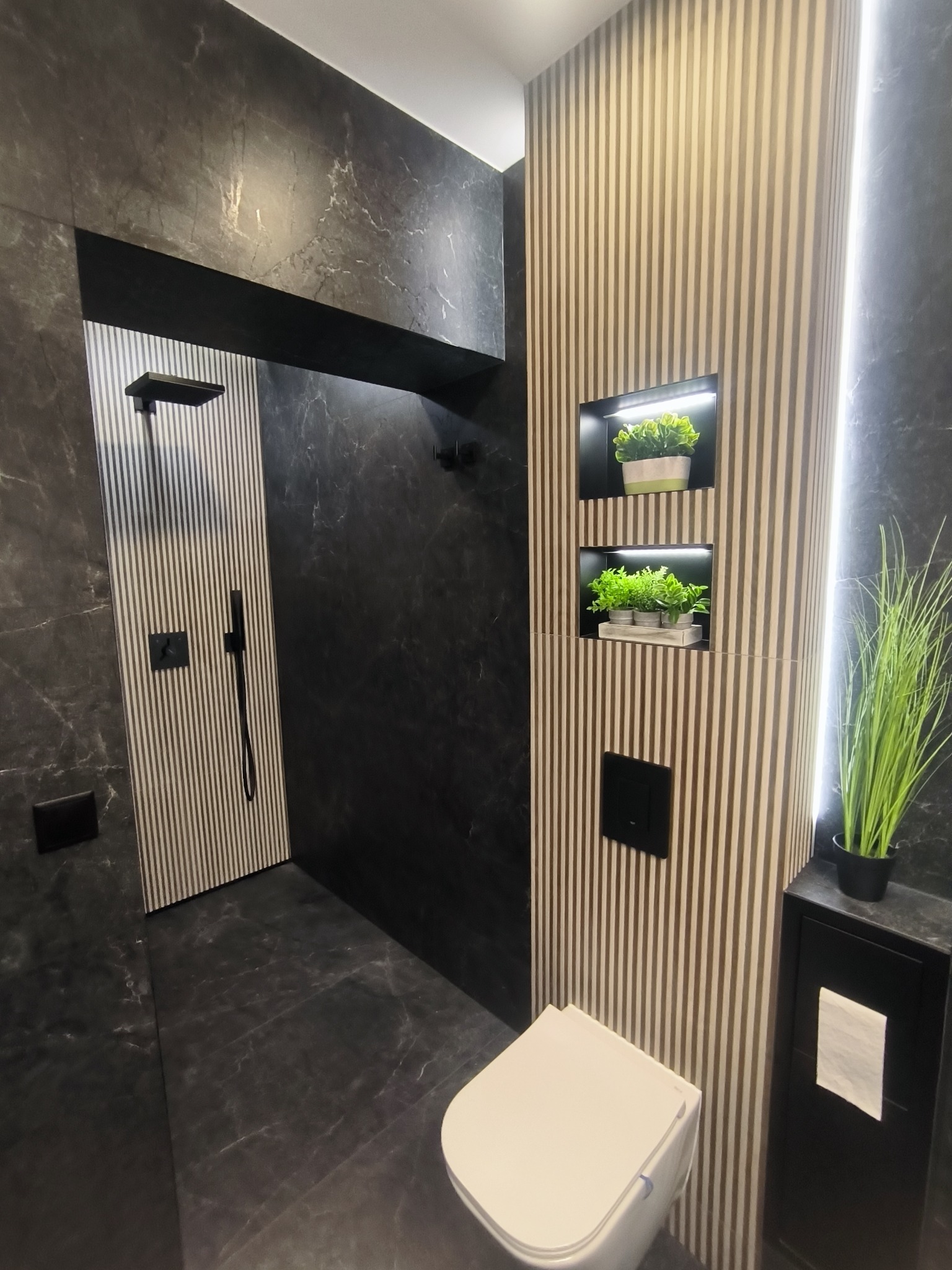 Nowoczesna łazienka z czarnymi marmurowymi płytkami, kabiną prysznicową typu walk-in i ścianą z pionowymi panelami z wnękami na rośliny. Minimalistyczny design.