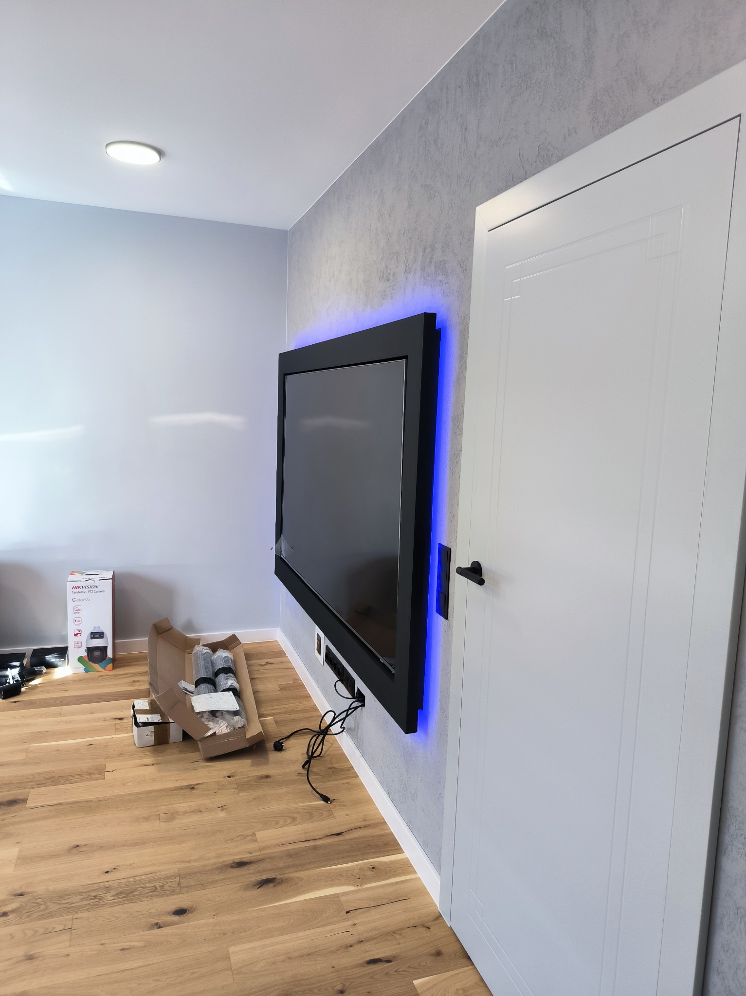 Telewizor z podświetleniem LED na szarej ścianie, obok białe drzwi. Na podłodze panele i karton z akcesoriami. Nowoczesne wykończenie wnętrza.