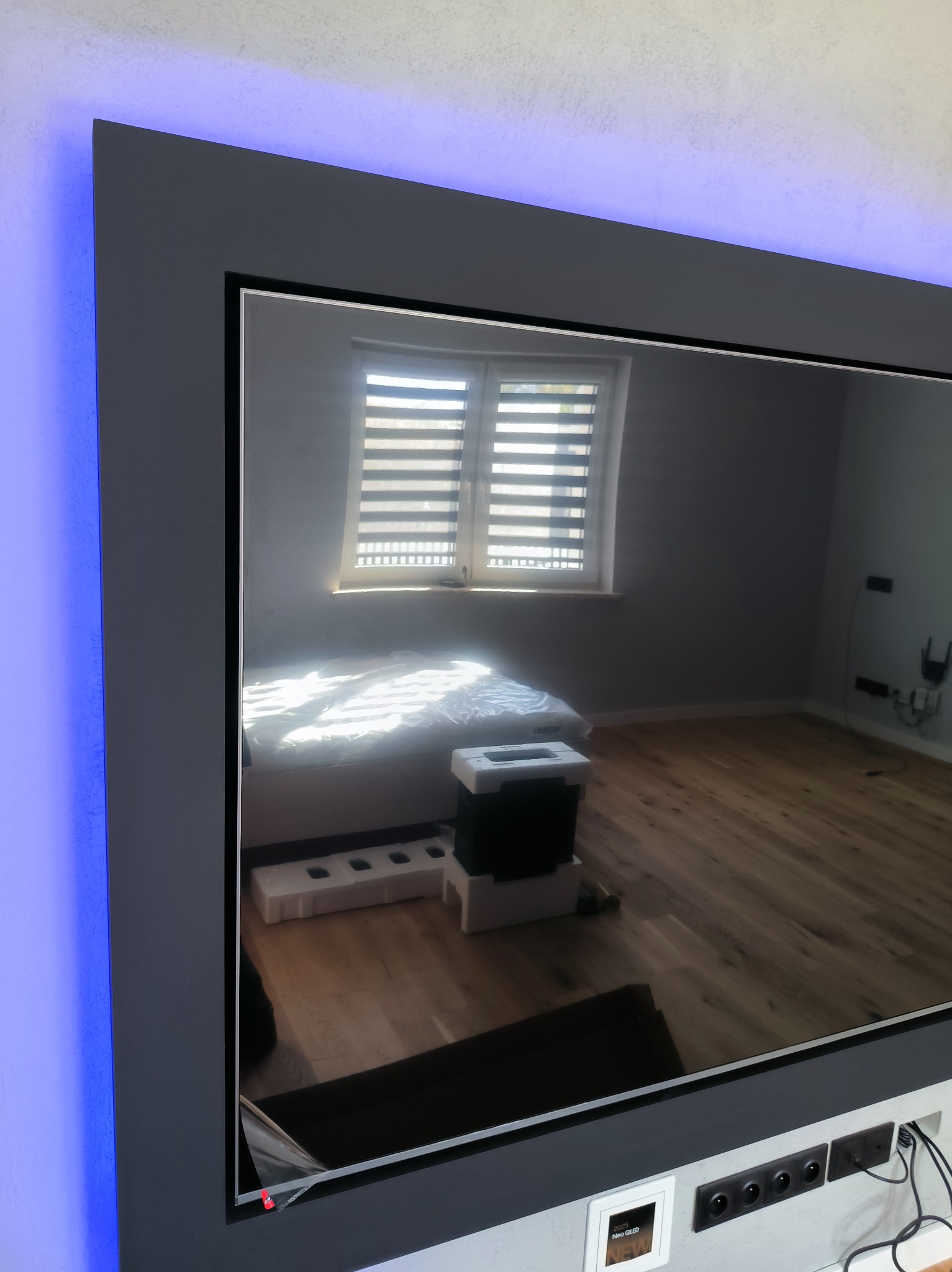 Telewizor Neo QLED w obudowie z podświetleniem LED, odbijający wnętrze pokoju z drewnianą podłogą i oknem z żaluzjami. Widoczne gniazdka elektryczne.