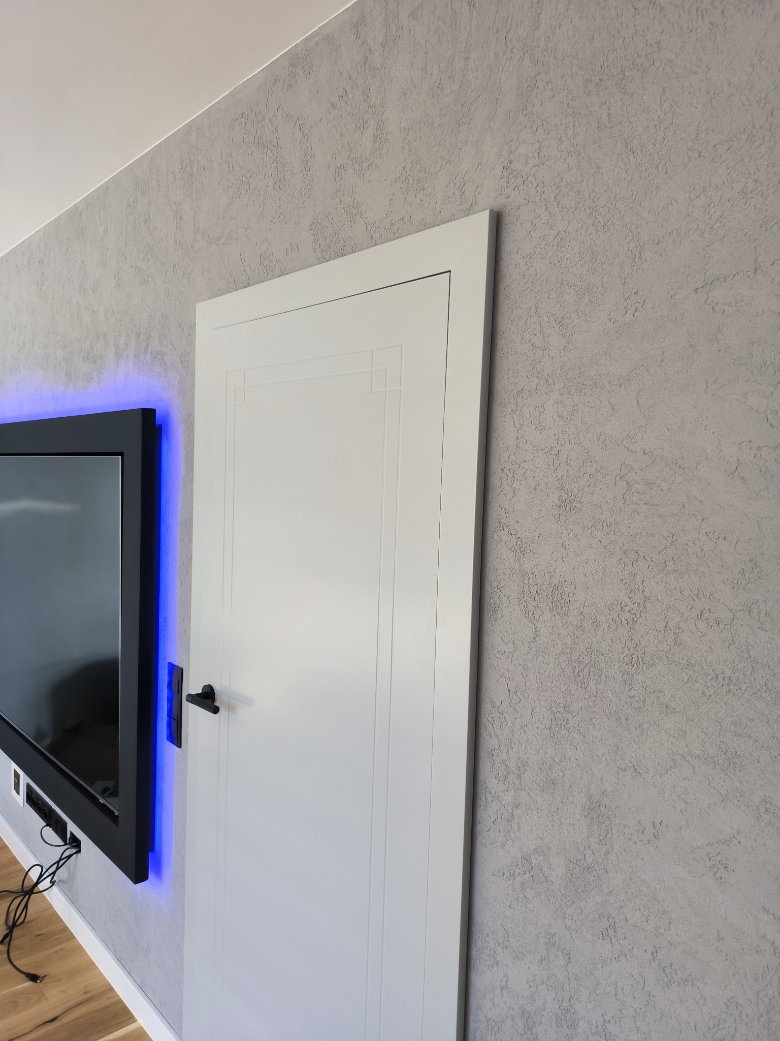 Szara ściana z fakturą, białe drzwi z czarną klamką i telewizor z niebieskim podświetleniem. Nowoczesne wykończenie wnętrza.