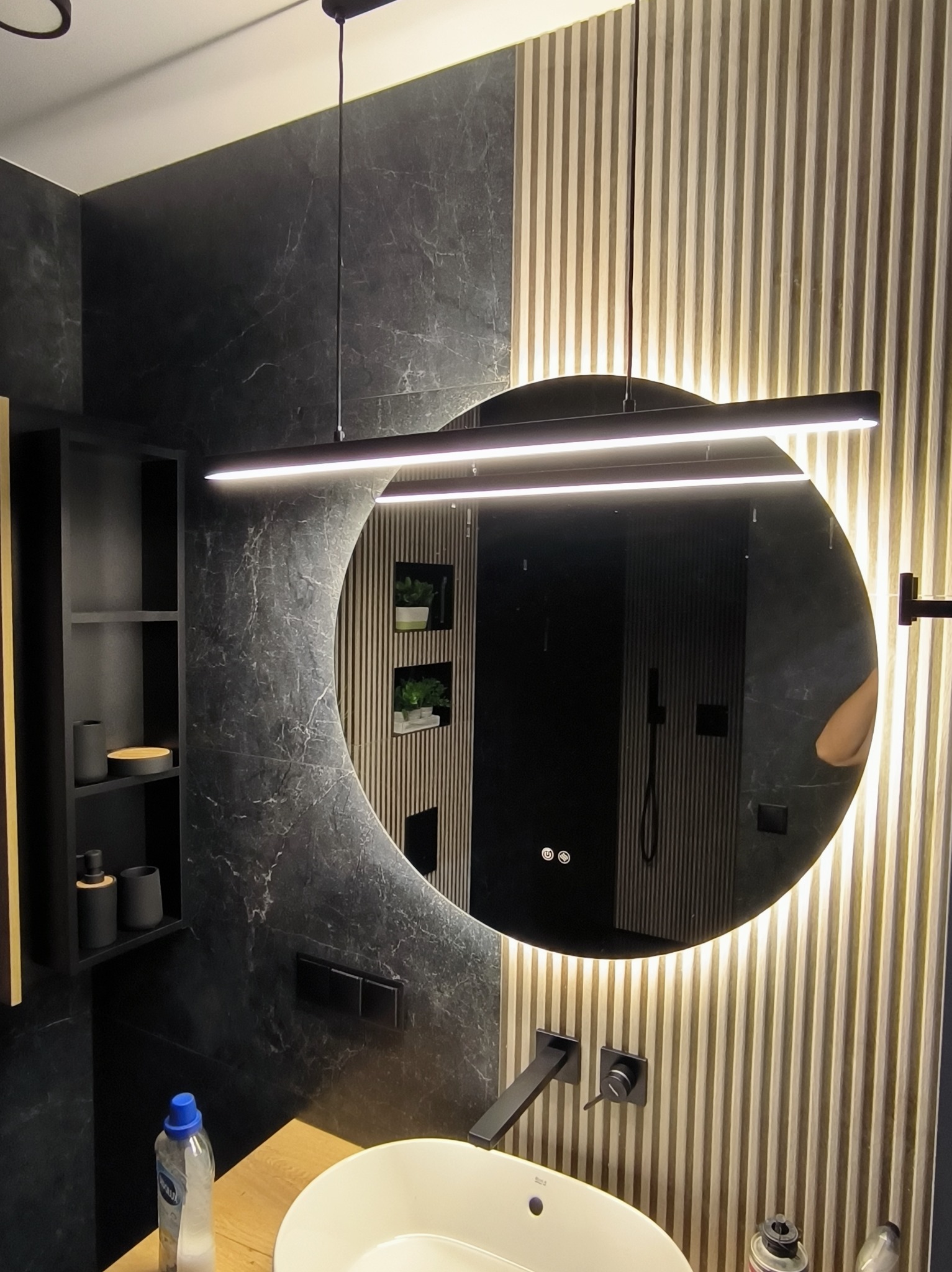 Nowoczesna łazienka z okrągłym lustrem LED, czarną armaturą i drewnianymi panelami ściennymi. Minimalistyczny design z akcentami marmuru i subtelnym oświetleniem.
