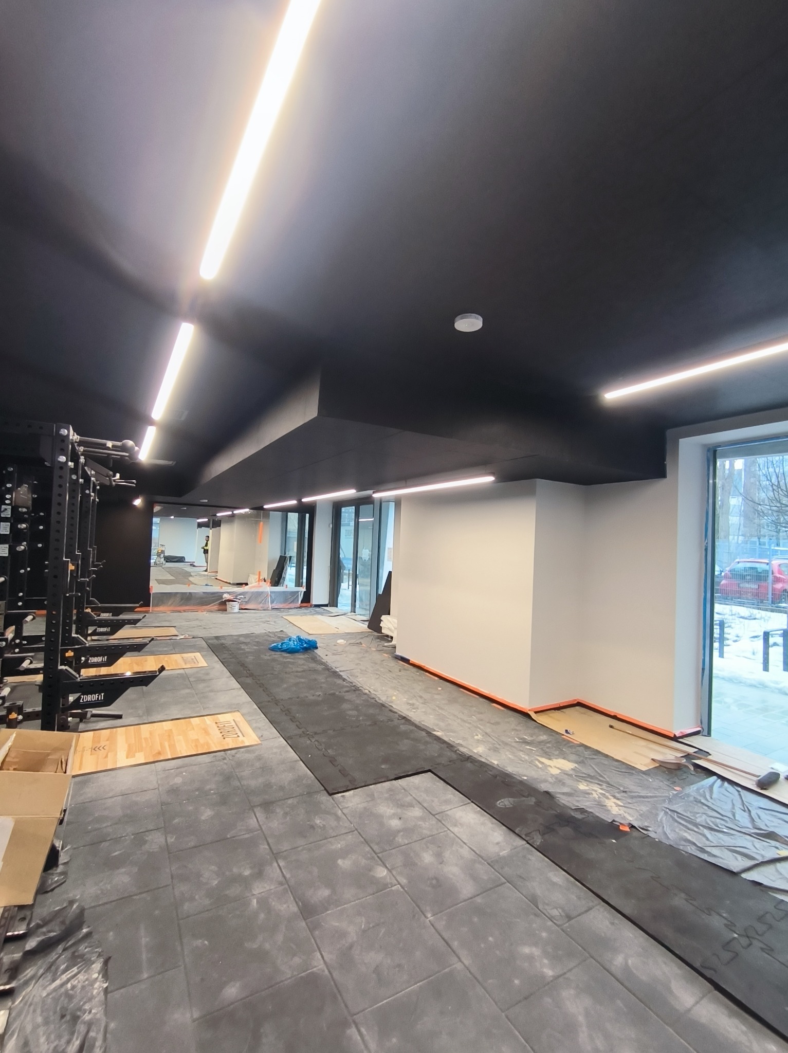 Montaż podłogi w siłowni: szare płytki, czarny sufit z oświetleniem LED, sprzęt do ćwiczeń. Widoczne etapy prac wykończeniowych, częściowo zabezpieczona podłoga.