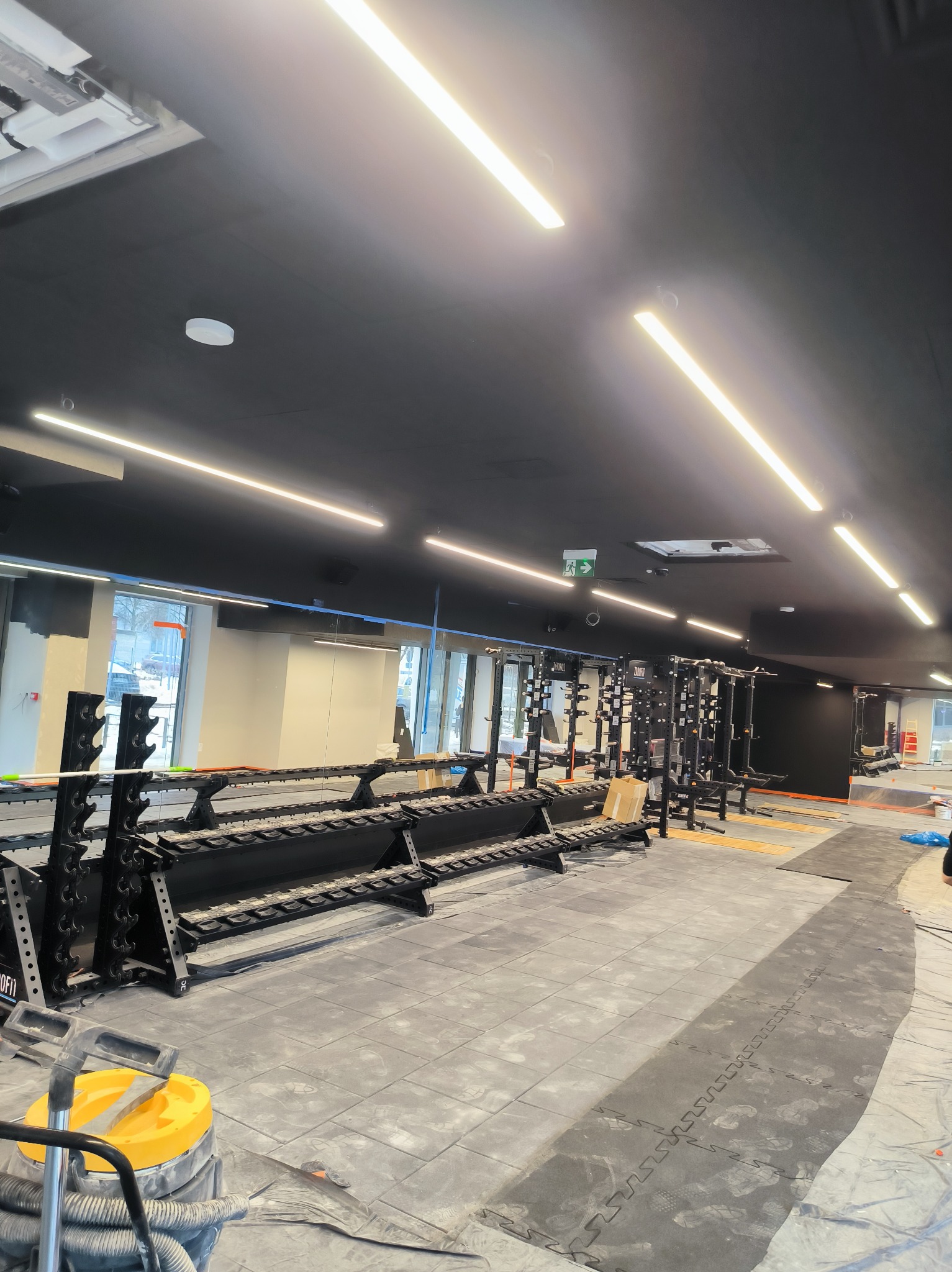 Montaż podłogi w nowej siłowni: gumowe puzzle na podłożu, widoczne regały na ciężary i sprzęt do ćwiczeń. Oświetlenie LED na czarnym suficie.
