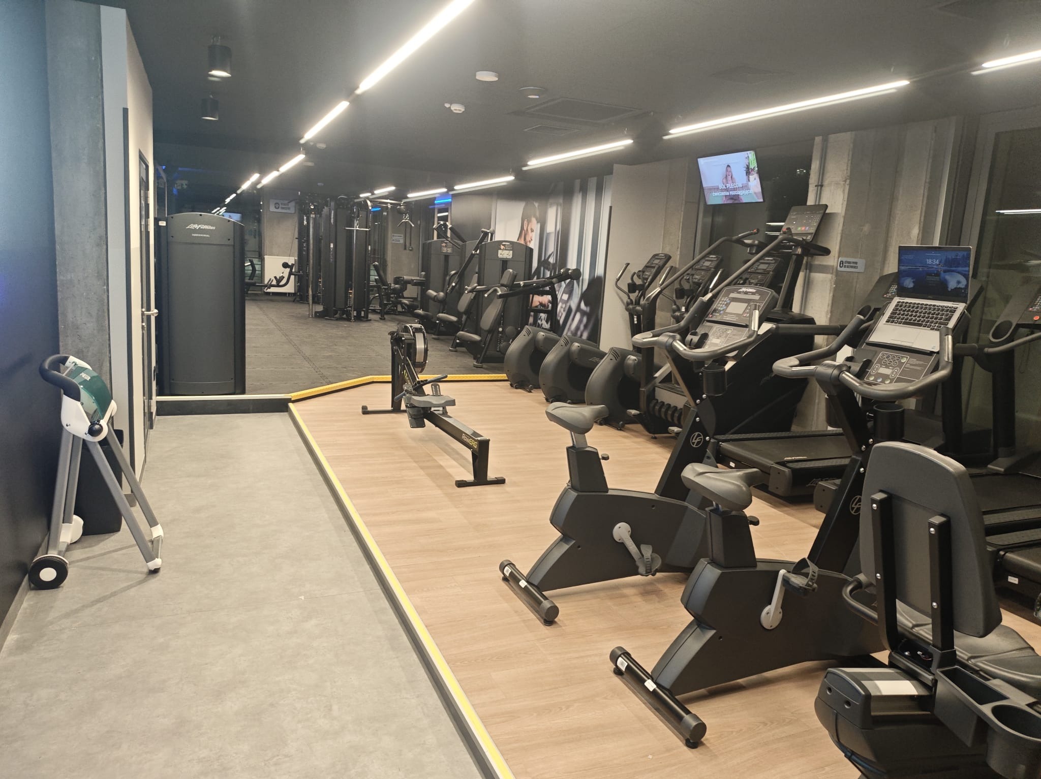 Nowoczesna siłownia z różnorodnym sprzętem: bieżnie z laptopem, rowery stacjonarne, wioślarz i maszyny siłowe. Jasne oświetlenie, betonowe akcenty na ścianach.