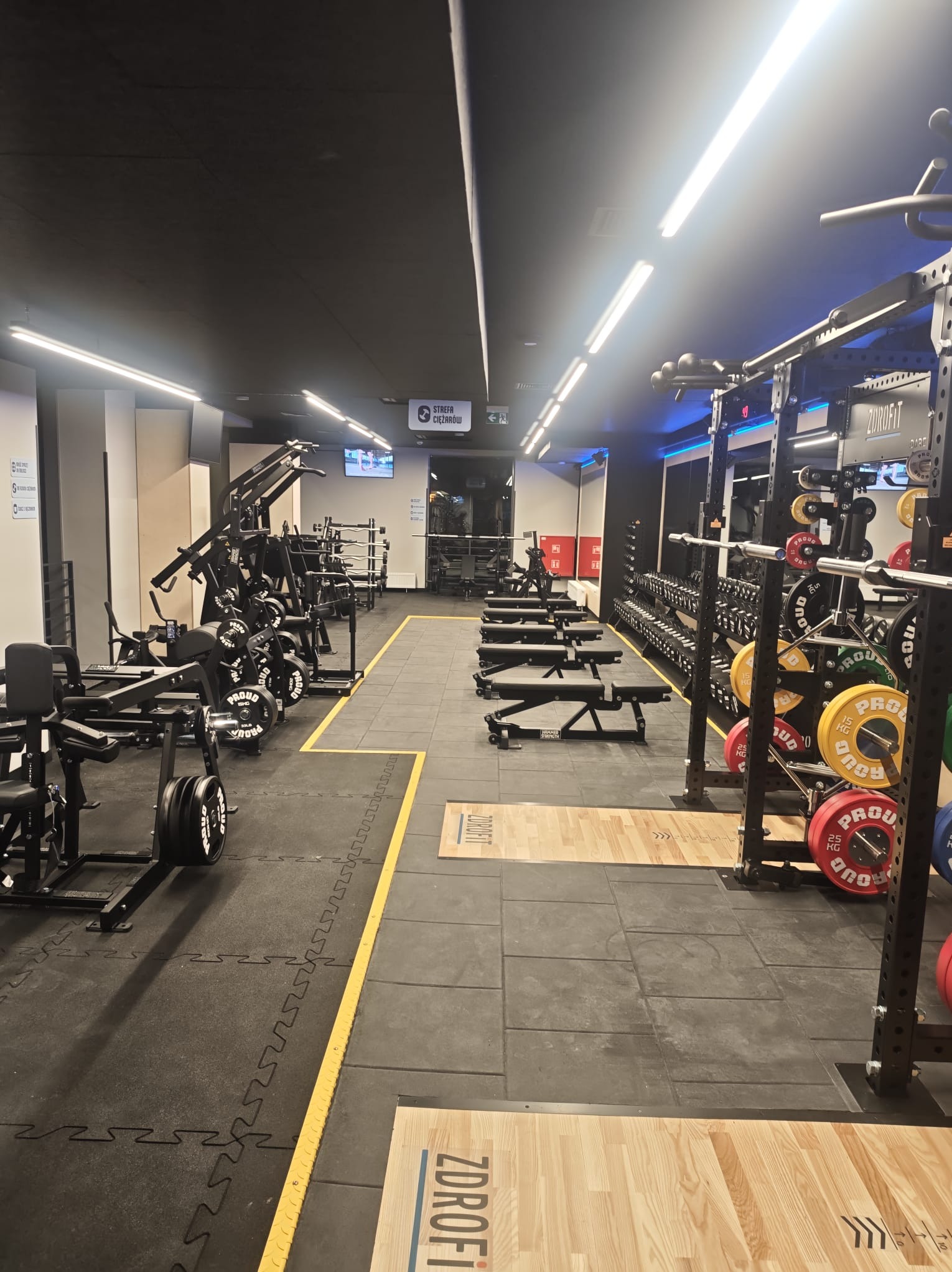 Nowoczesna siłownia Zdrofit z czarną podłogą i sufitem, rzędy maszyn do ćwiczeń, sztangi i obciążenia. Jasne oświetlenie LED. Widok w głąb sali.