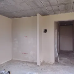 Surowe wnętrze po nałożeniu jasnego tynku, widoczne gniazdka elektryczne, betonowy sufit, drabina oparta o ścianę w przejściu, przygotowane do dalszych prac wykończeniowych.