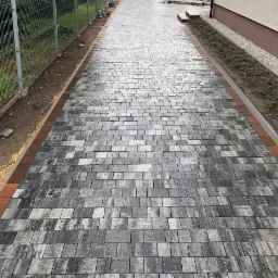 Długa, wybrukowana ścieżka z szarej kostki o nieregularnej fakturze, obramowana czerwoną kostką, prowadząca wzdłuż ogrodzenia i budynku z rabatą.