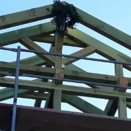 Drewniana konstrukcja więźby dachowej z wieńcem zawieszonym na szczycie, widoczna z poziomu rusztowania.