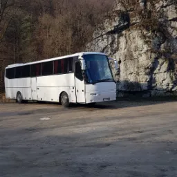 Biały autokar turystyczny zaparkowany na żwirowym placu, w tle skała z pomnikiem i drzewa bez liści.