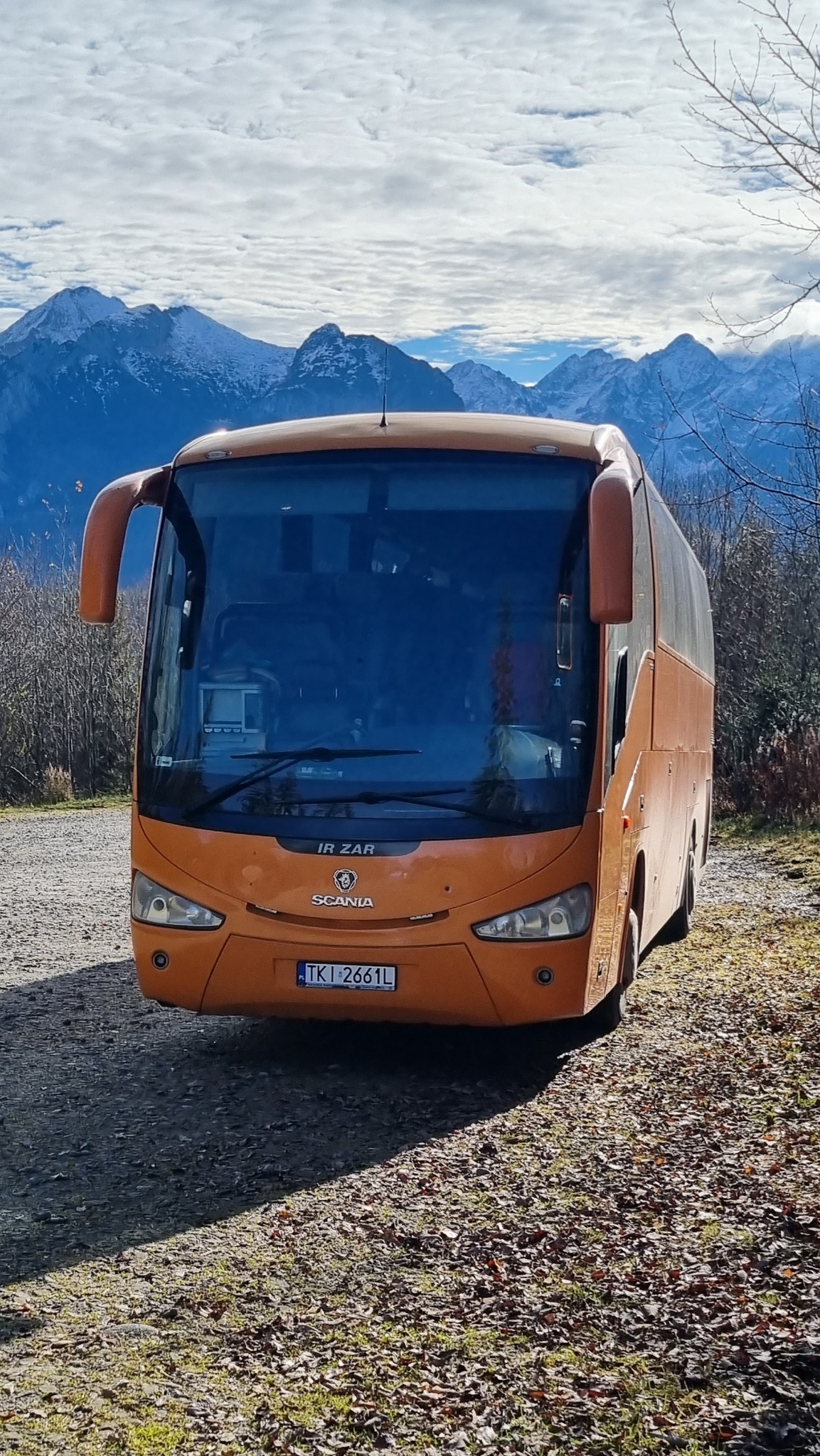 Pomarańczowy autokar Scania IR ZAR z numerem rejestracyjnym TKI 2661L na tle górskiego krajobrazu z ośnieżonymi szczytami.