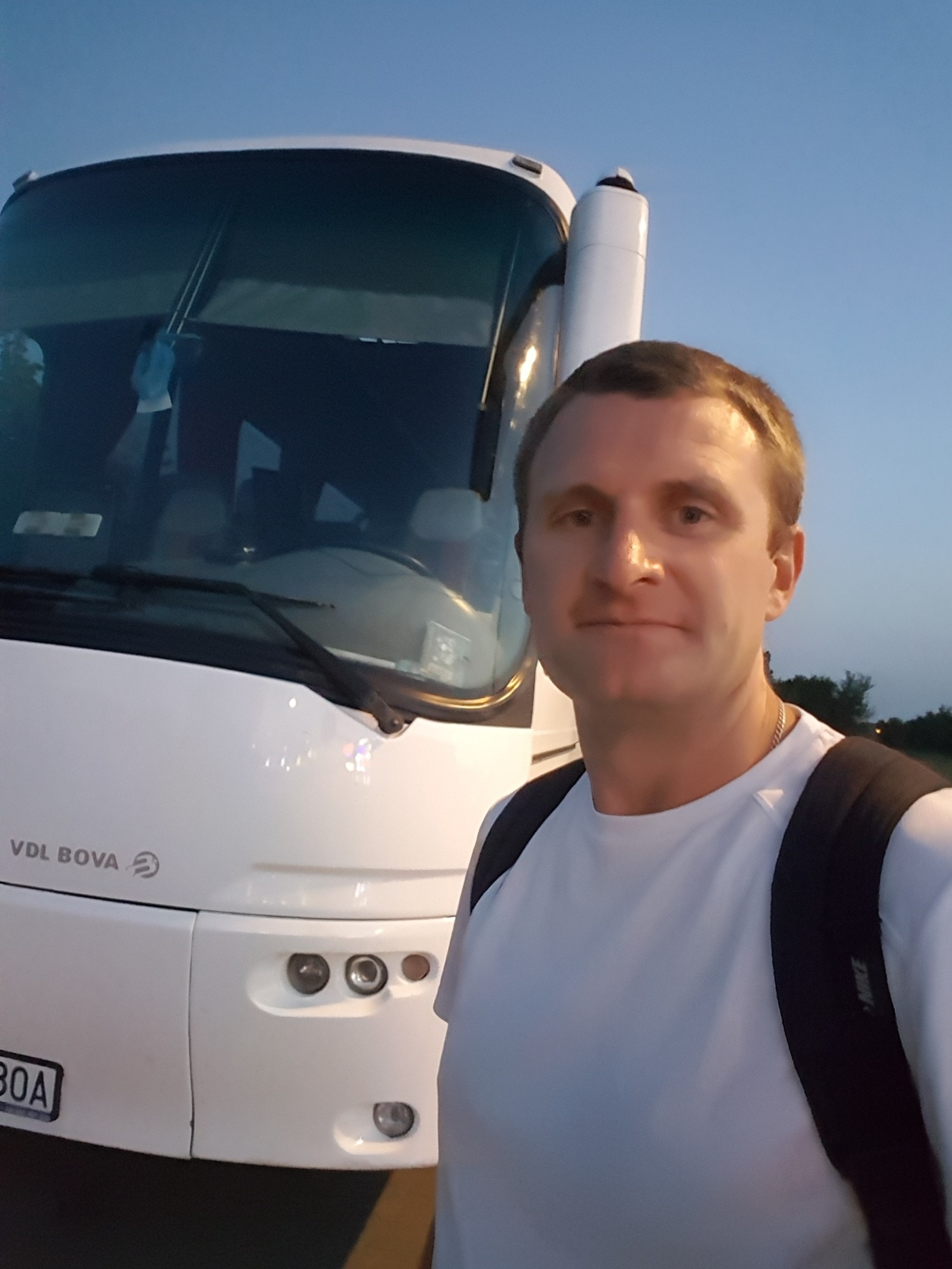 Mężczyzna z plecakiem pozuje na tle białego autobusu VDL BOVA z widoczną tablicą rejestracyjną i elementami wyposażenia.