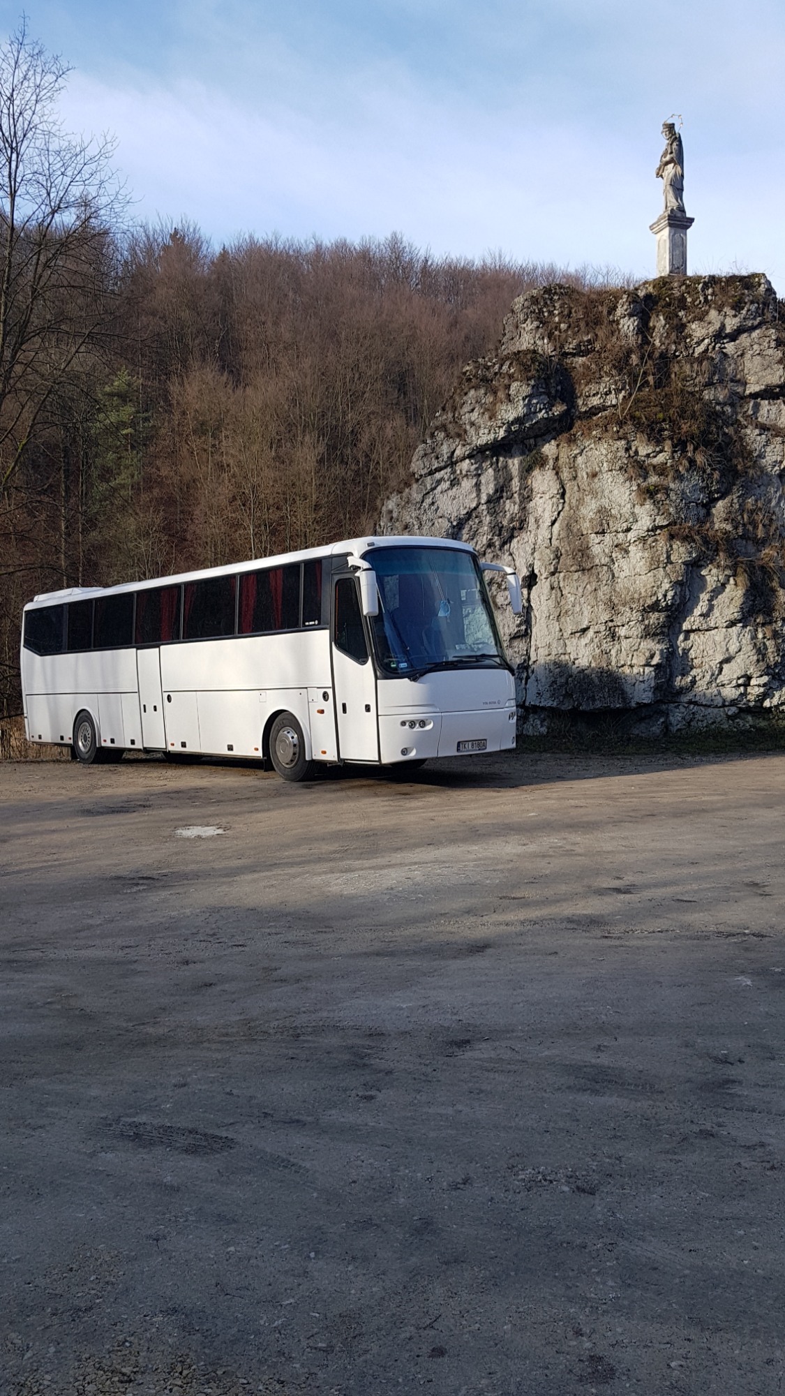 Biały autokar turystyczny zaparkowany na żwirowym placu, w tle skała z pomnikiem i drzewa bez liści.