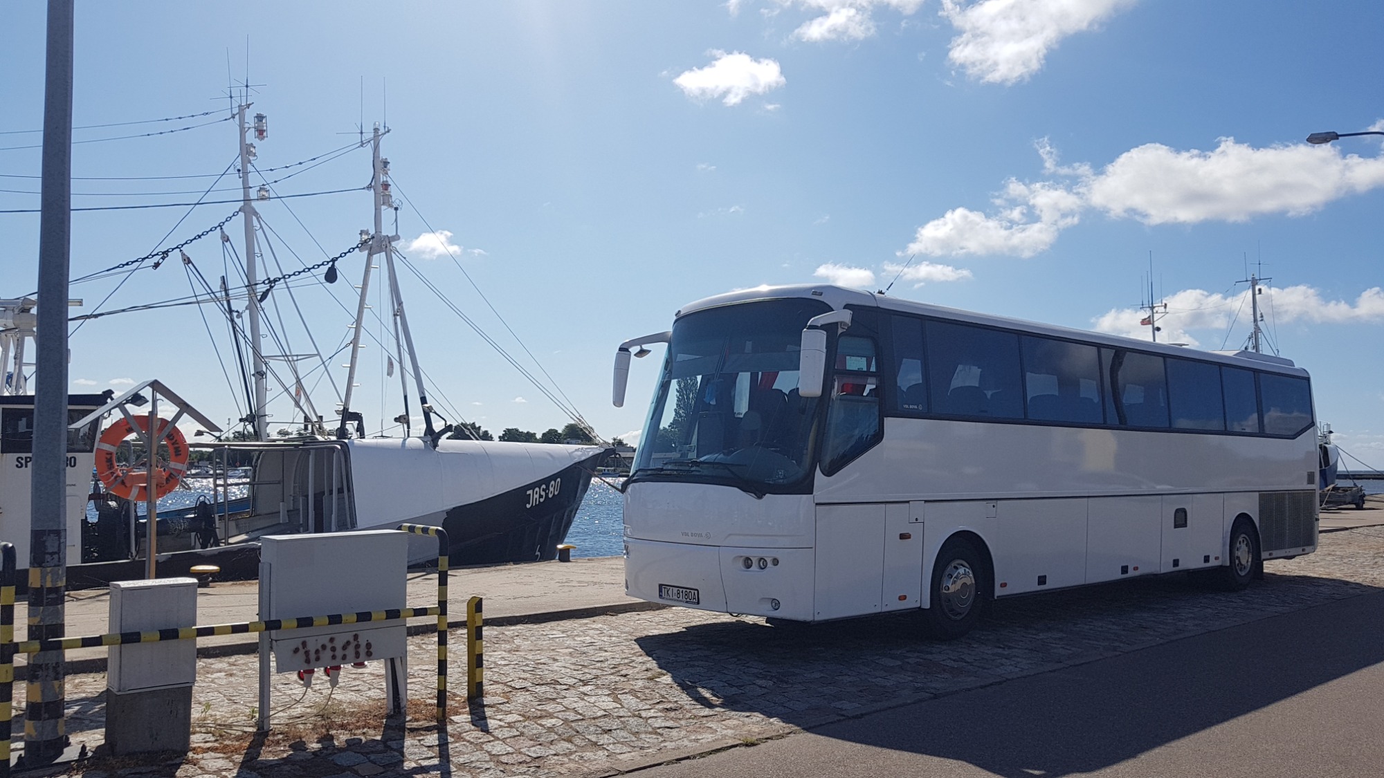 Biały autokar turystyczny na tle portu rybackiego w Górnie, z widocznymi masztami i łodziami rybackimi. Na pierwszym planie kostka brukowa.