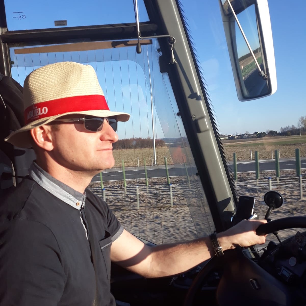 Kierowca autobusu w kapeluszu z czerwoną wstążką, prowadzący pojazd w słoneczny dzień, widok na krajobraz przez szybę.