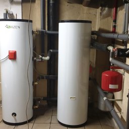 AMS Solar - Dwa białe, cylindryczne zasobniki ciepłej wody użytkowej marki Shizen w kotłowni, z widocznymi rurami, zaworami i osprzętem hydraulicznym.