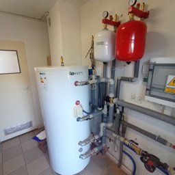 AMS Solar - Biały zasobnik ciepłej wody użytkowej marki Shizen z podłączonymi rurami, manometrami i zbiornikami wyrównawczymi w kolorze białym i czerwonym, zamontowany w pomieszczeniu z płytkami na podłodze.