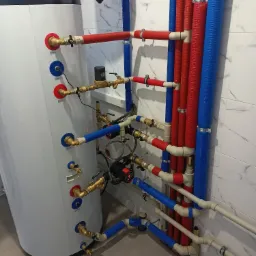 Instalacja kotła grzewczego z licznymi rurami w kolorach czerwonym i niebieskim, widoczne zawory i pompy obiegowe, tło stanowi marmurkowa ściana.