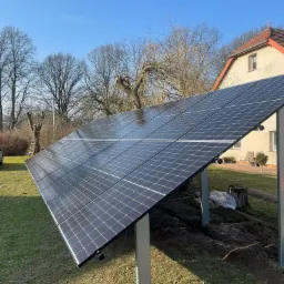Panele słoneczne na trawniku przed domem z czerwoną dachówką w słoneczny dzień. Widoczne drzewa bez liści i drabina oparta o drzewo w tle.