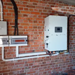 Biała skrzynka z wyświetlaczem i instalacją elektryczną na ceglanej ścianie. Widoczne przewody i rury instalacyjne. Elementy systemu zasilania.