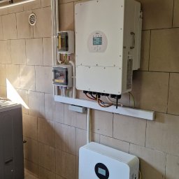 AMS FULL SERWIS Marcin Kaczmarek - Zainstalowany biały inwerter Deye i akumulator Lithtech na ścianie z beżowej cegły w garażu. Widoczne przewody i skrzynki elektryczne. Realizacja instalacji fotowoltaicznej.