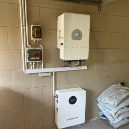 AMS FULL SERWIS Marcin Kaczmarek - Kompletna instalacja z białym inwerterem i magazynem energii LITH TECH na beżowej ścianie z bloków. Widoczne kable, rury i skrzynki elektryczne. Podłoga wyłożona płytkami.