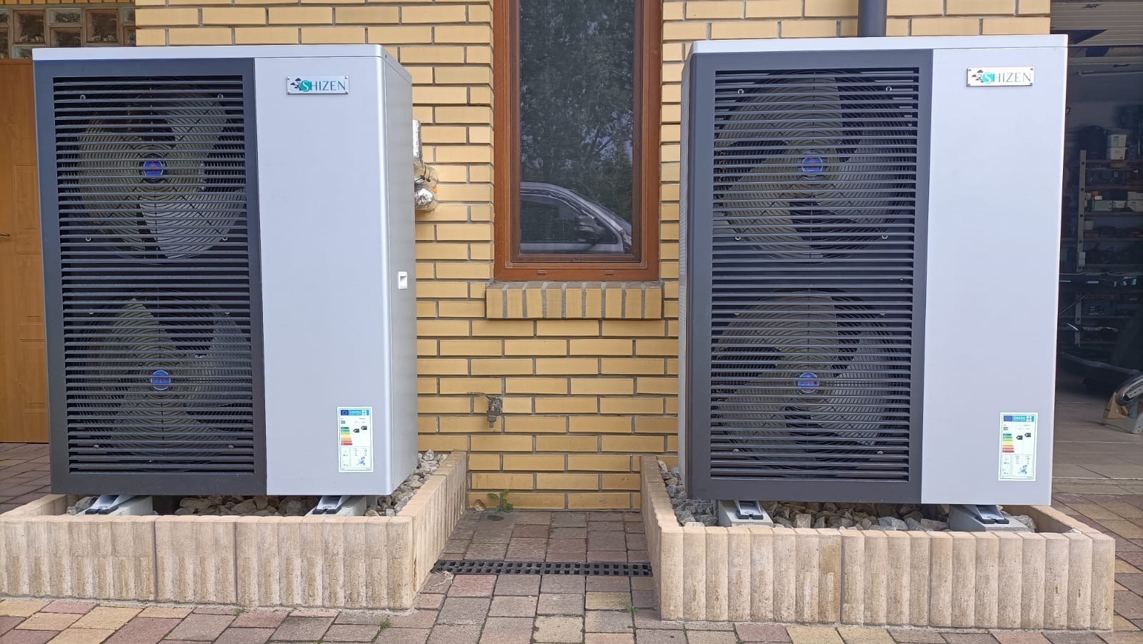 Dwie szare pompy ciepła marki Shizen ustawione na podwyższeniach z kostki betonowej przed ceglanym budynkiem z oknem, widoczna kratka ściekowa.