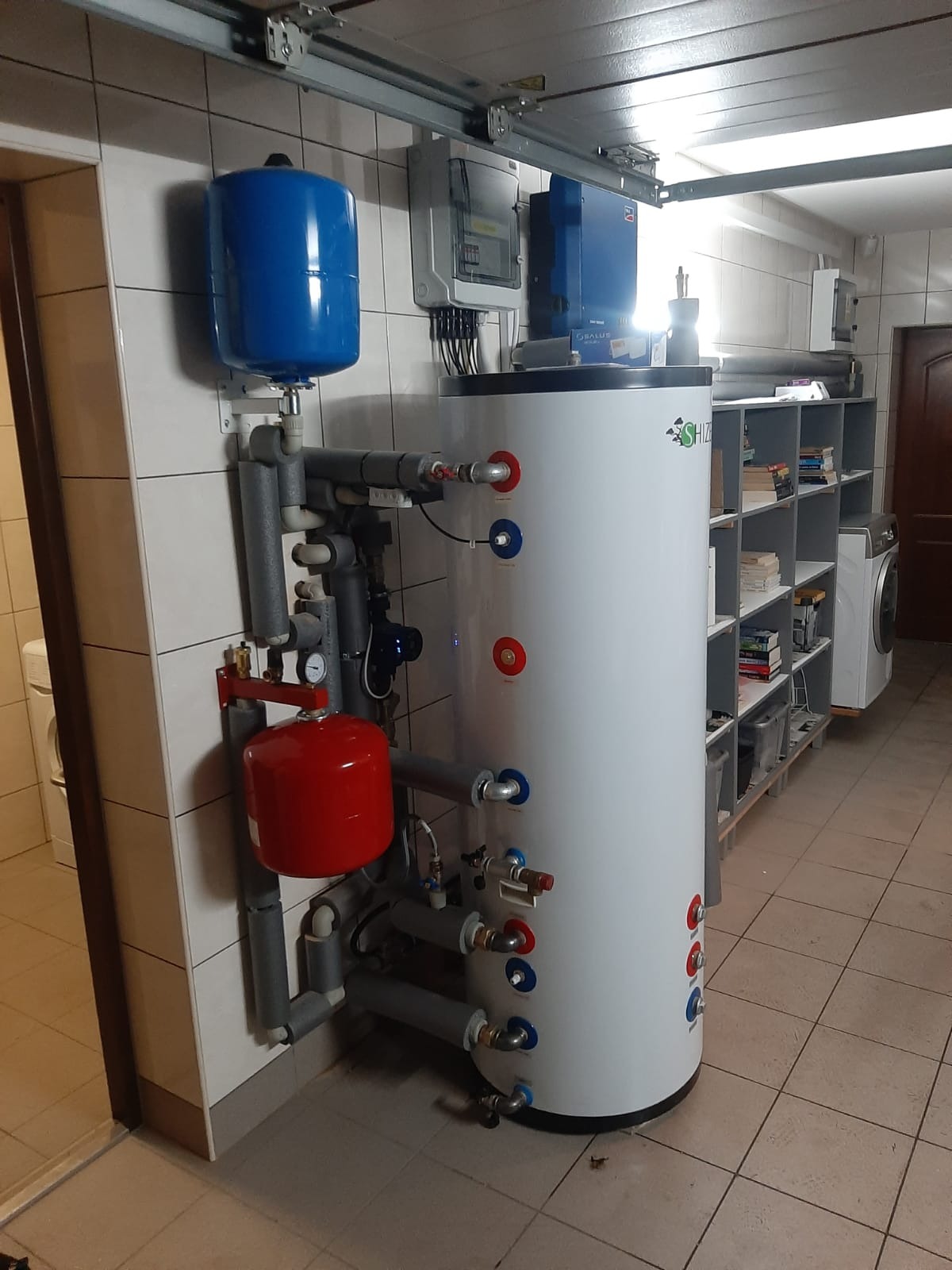 Bojler akumulacyjny z osprzętem hydraulicznym w kotłowni, widoczne zbiorniki wyrównawcze w kolorach niebieskim i czerwonym, instalacja w szarych izolacjach.