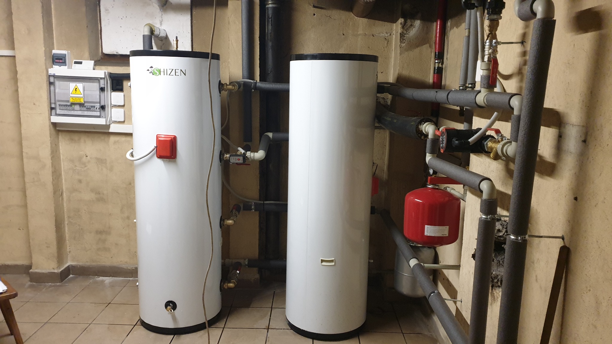 Dwa białe, cylindryczne zasobniki ciepłej wody użytkowej marki Shizen w kotłowni, z widocznymi rurami, zaworami i osprzętem hydraulicznym.