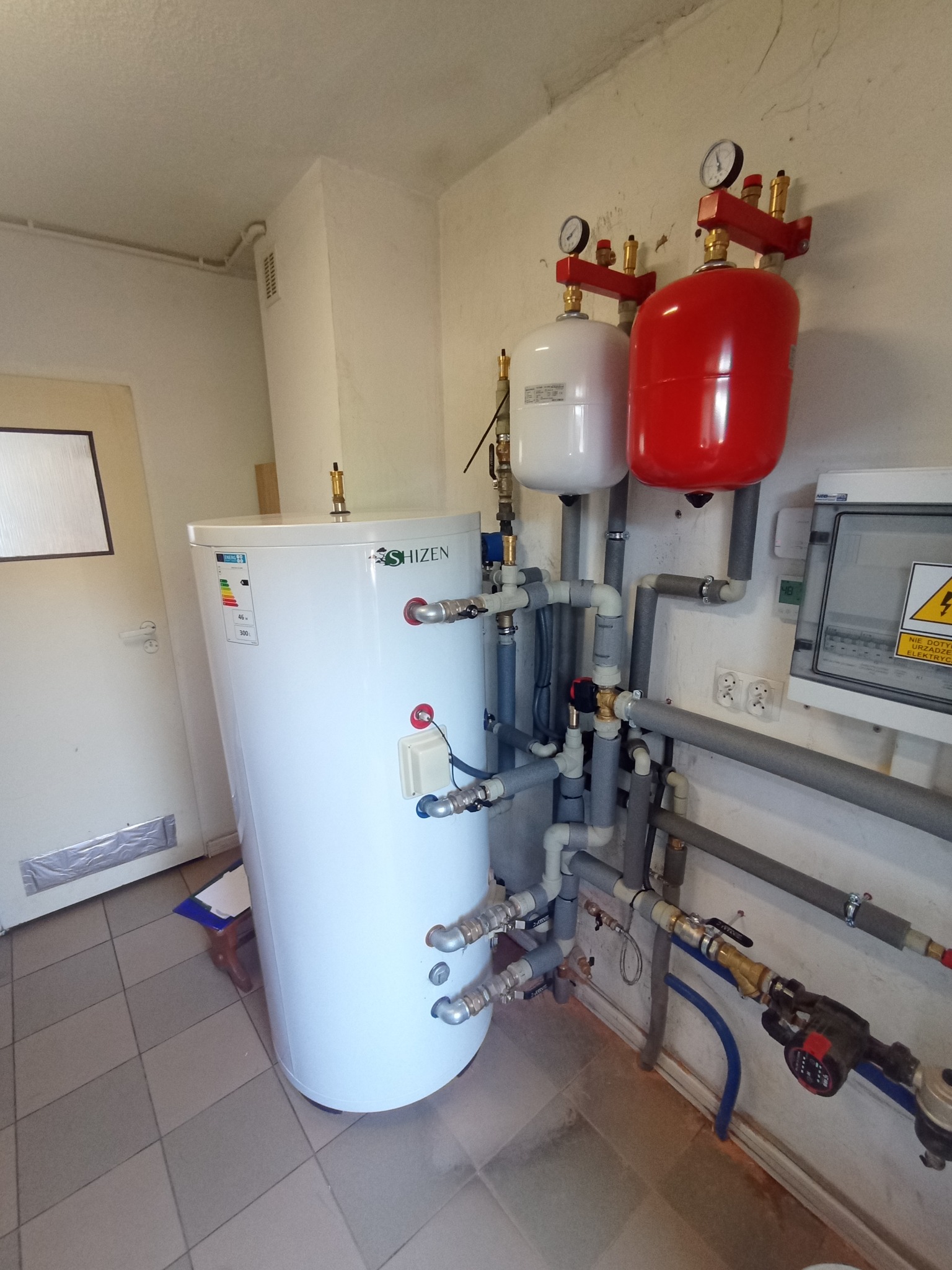 Biały zasobnik ciepłej wody użytkowej marki Shizen z podłączonymi rurami, manometrami i zbiornikami wyrównawczymi w kolorze białym i czerwonym, zamontowany w pomieszczeniu z płytkami na podłodze.