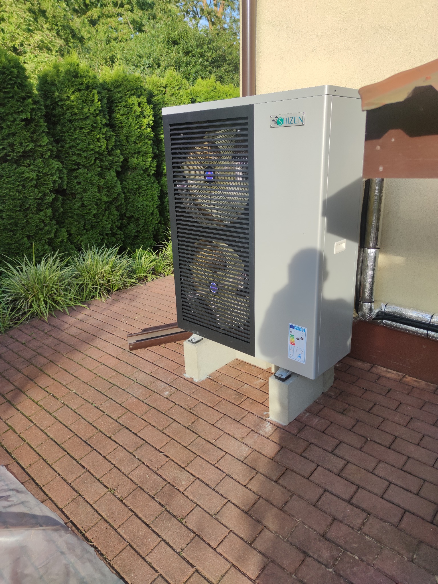 Szara pompa ciepła Shizen z dwoma wentylatorami, zamontowana na betonowych podstawach na ceglanym patio, z zielonymi tujami w tle.
