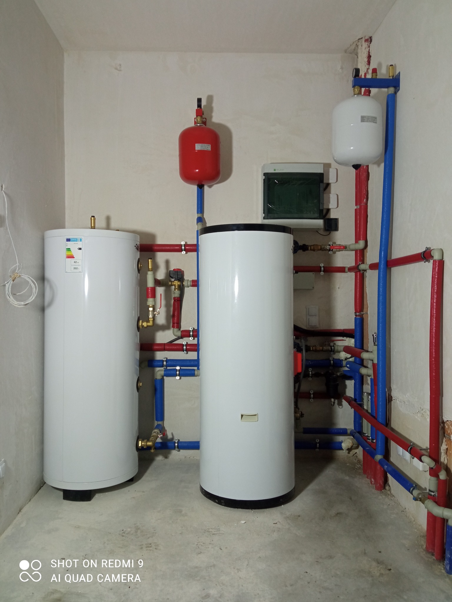 Dwa białe zbiorniki buforowe z czerwonymi i niebieskimi rurami w surowym pomieszczeniu technicznym z widocznymi śladami prac wykończeniowych na ścianach.