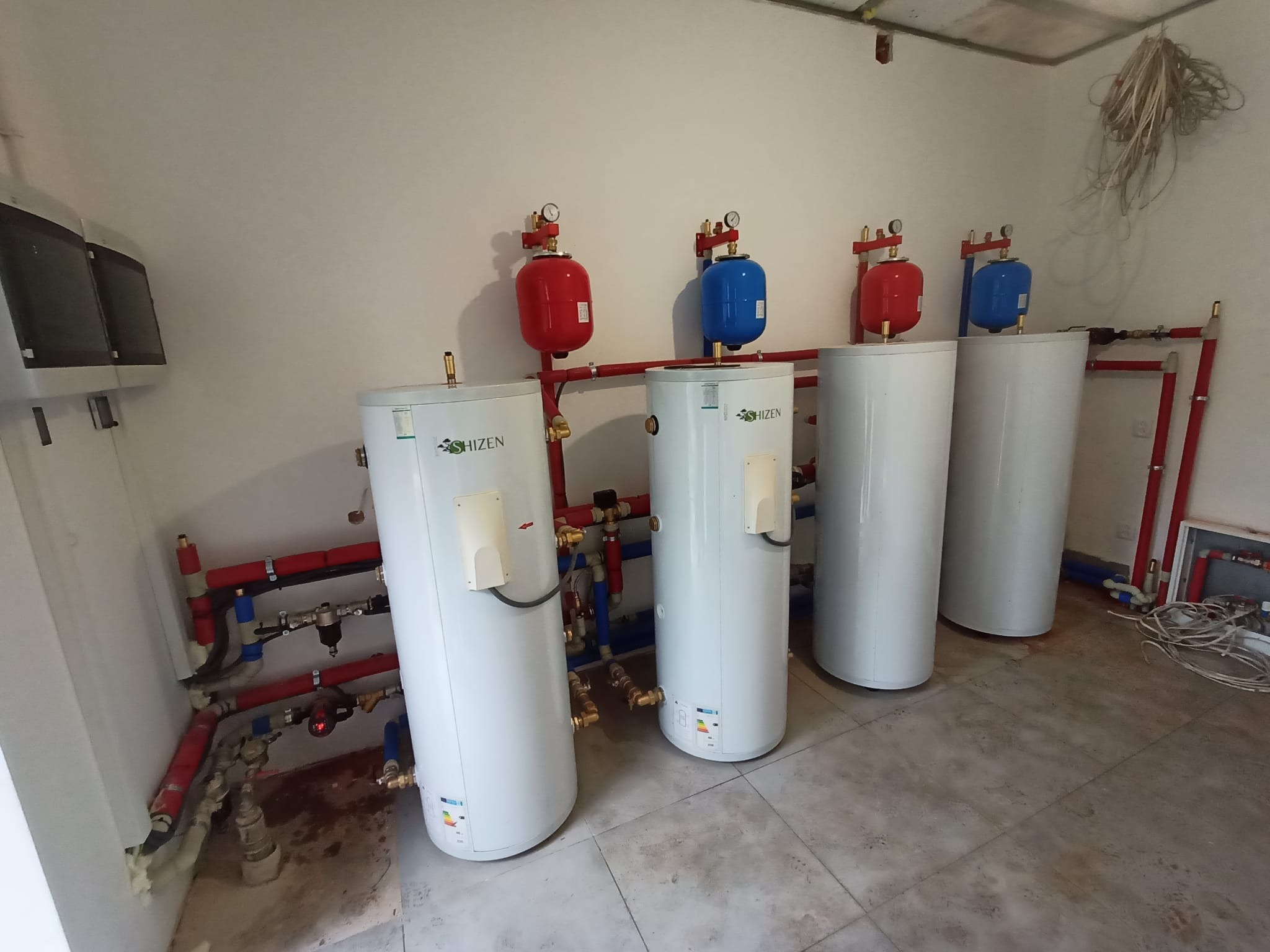 Instalacja czterech białych, cylindrycznych zbiorników z czerwonymi i niebieskimi naczyniami wzbiorczymi oraz rurami w pomieszczeniu z szarą podłogą z płytek.