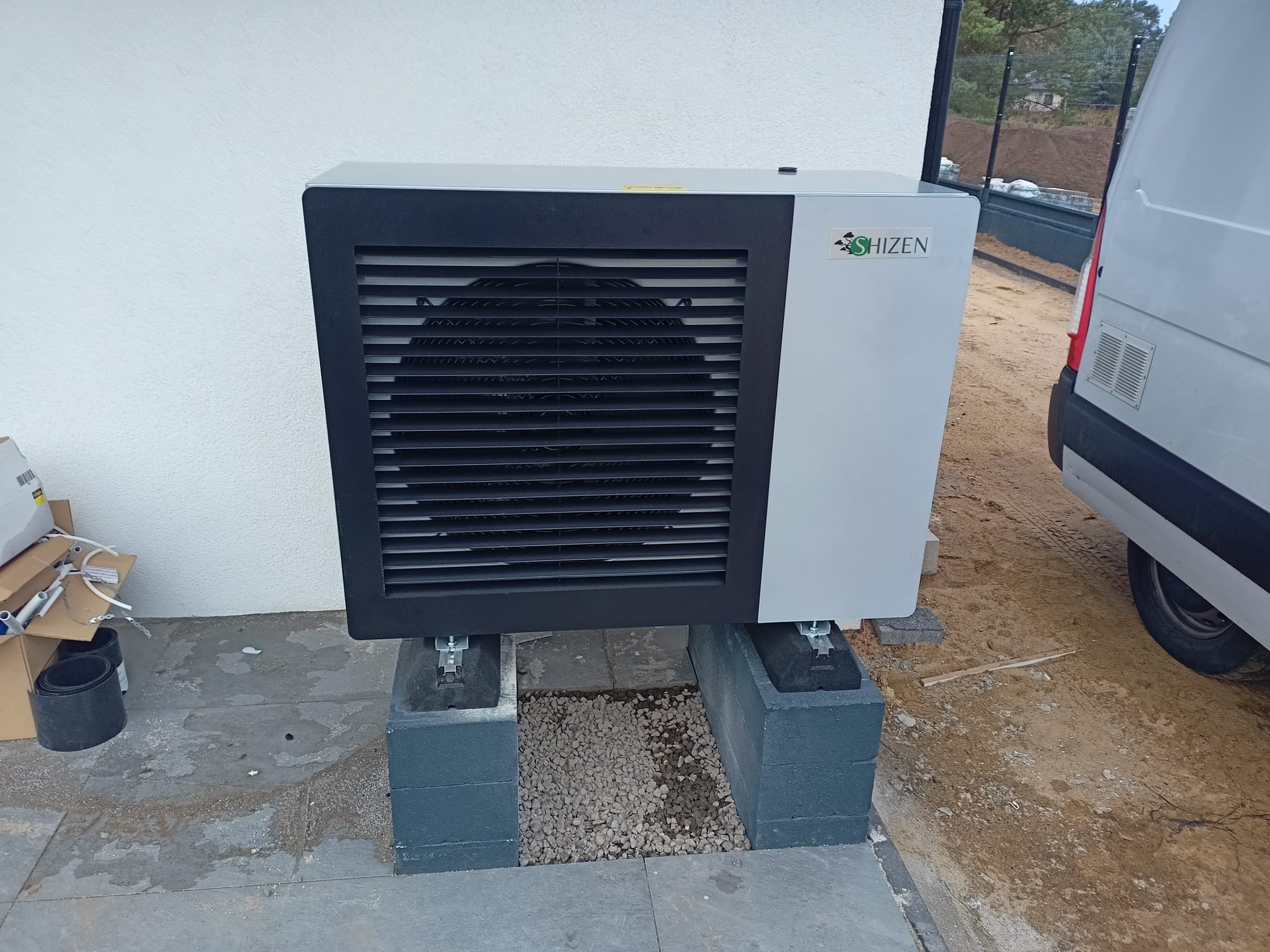 Zewnętrzna jednostka pompy ciepła marki Shizen, zamontowana na betonowych blokach, z widocznym fragmentem białego busa i przygotowanym podłożem gruntowym.