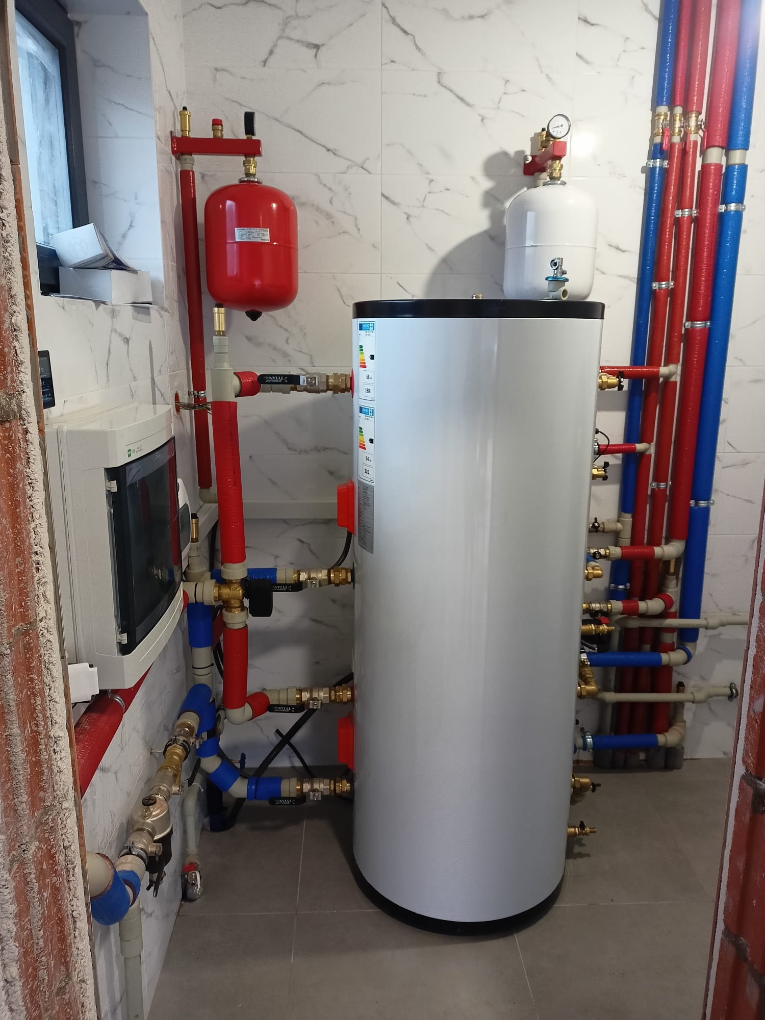 Biały, cylindryczny zbiornik buforowy ciepłej wody użytkowej z licznymi podłączeniami rur w kolorach czerwonym i niebieskim, zamontowany w pomieszczeniu z marmurowymi płytkami.