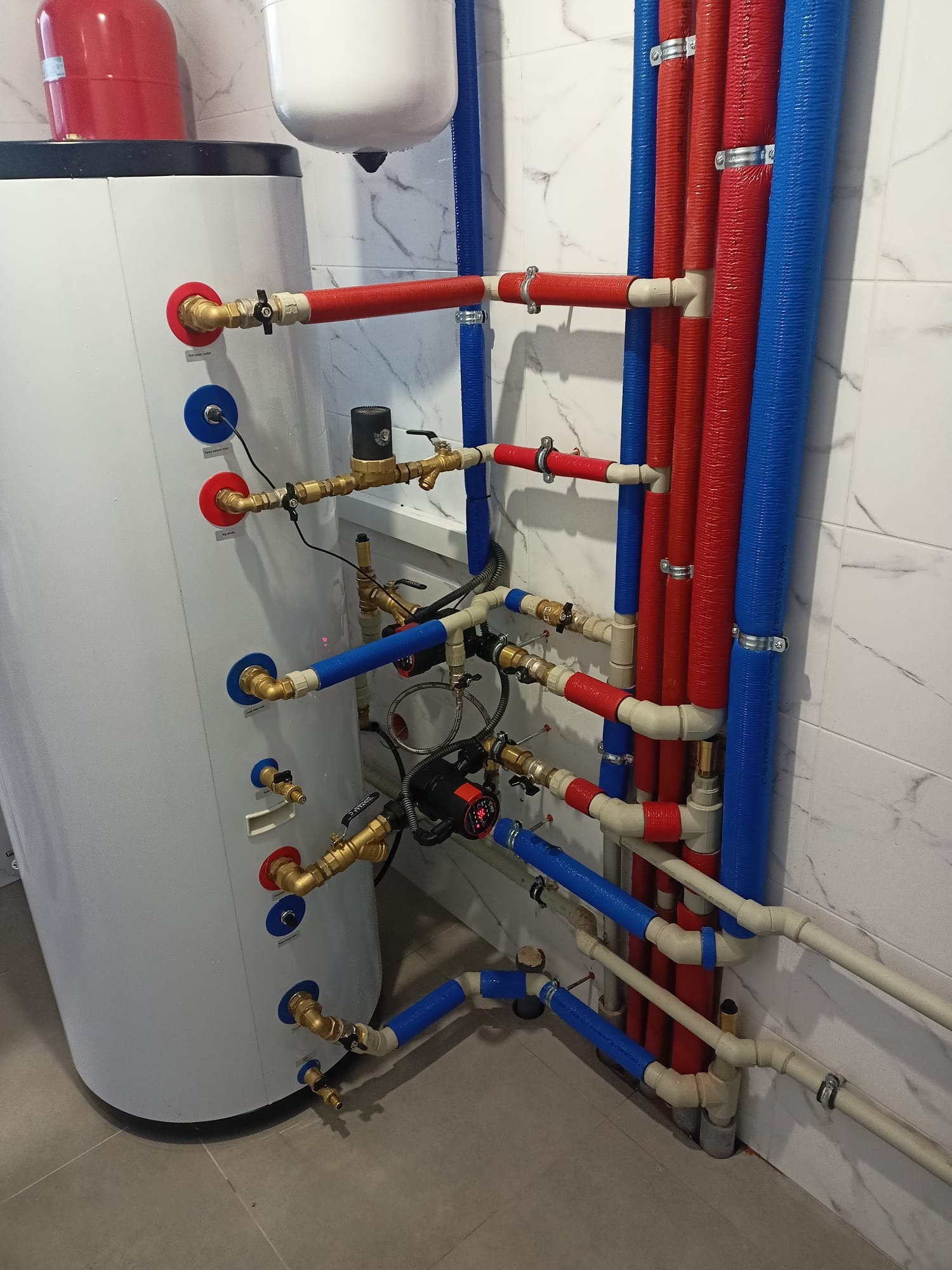 Instalacja kotła grzewczego z licznymi rurami w kolorach czerwonym i niebieskim, widoczne zawory i pompy obiegowe, tło stanowi marmurkowa ściana.