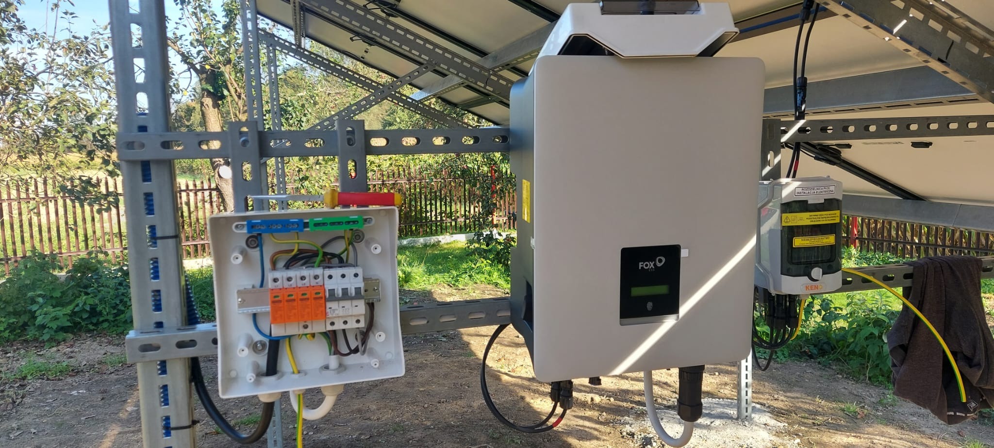 Instalacja paneli słonecznych: skrzynka elektryczna z bezpiecznikami, inwerter FOX ESS i moduł rozdzielczy na tle zieleni.