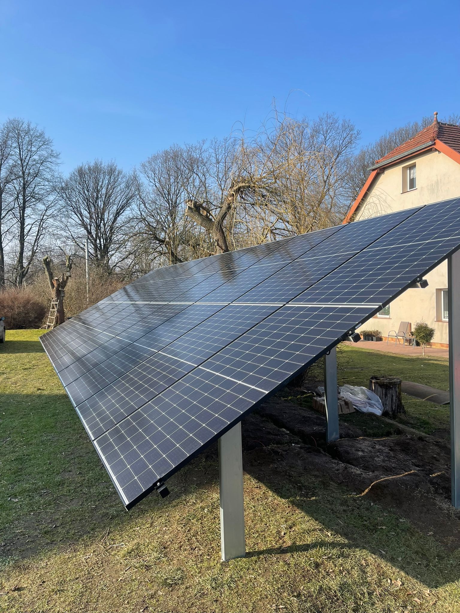 Panele słoneczne na trawniku przed domem z czerwoną dachówką w słoneczny dzień. Widoczne drzewa bez liści i drabina oparta o drzewo w tle.