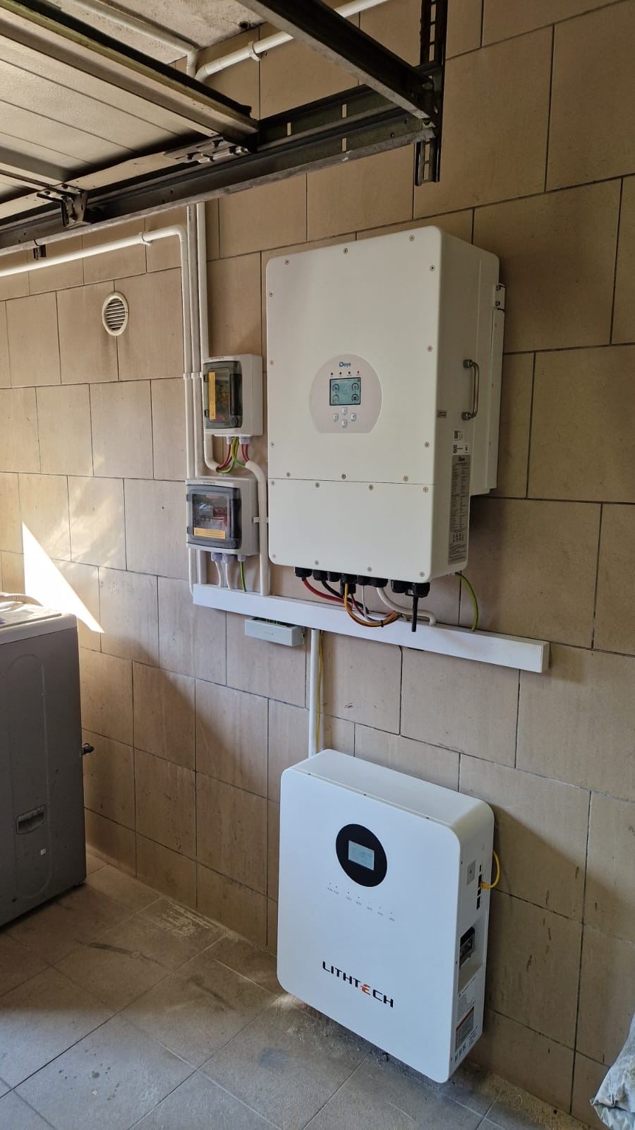 Zainstalowany biały inwerter Deye i akumulator Lithtech na ścianie z beżowej cegły w garażu. Widoczne przewody i skrzynki elektryczne. Realizacja instalacji fotowoltaicznej.
