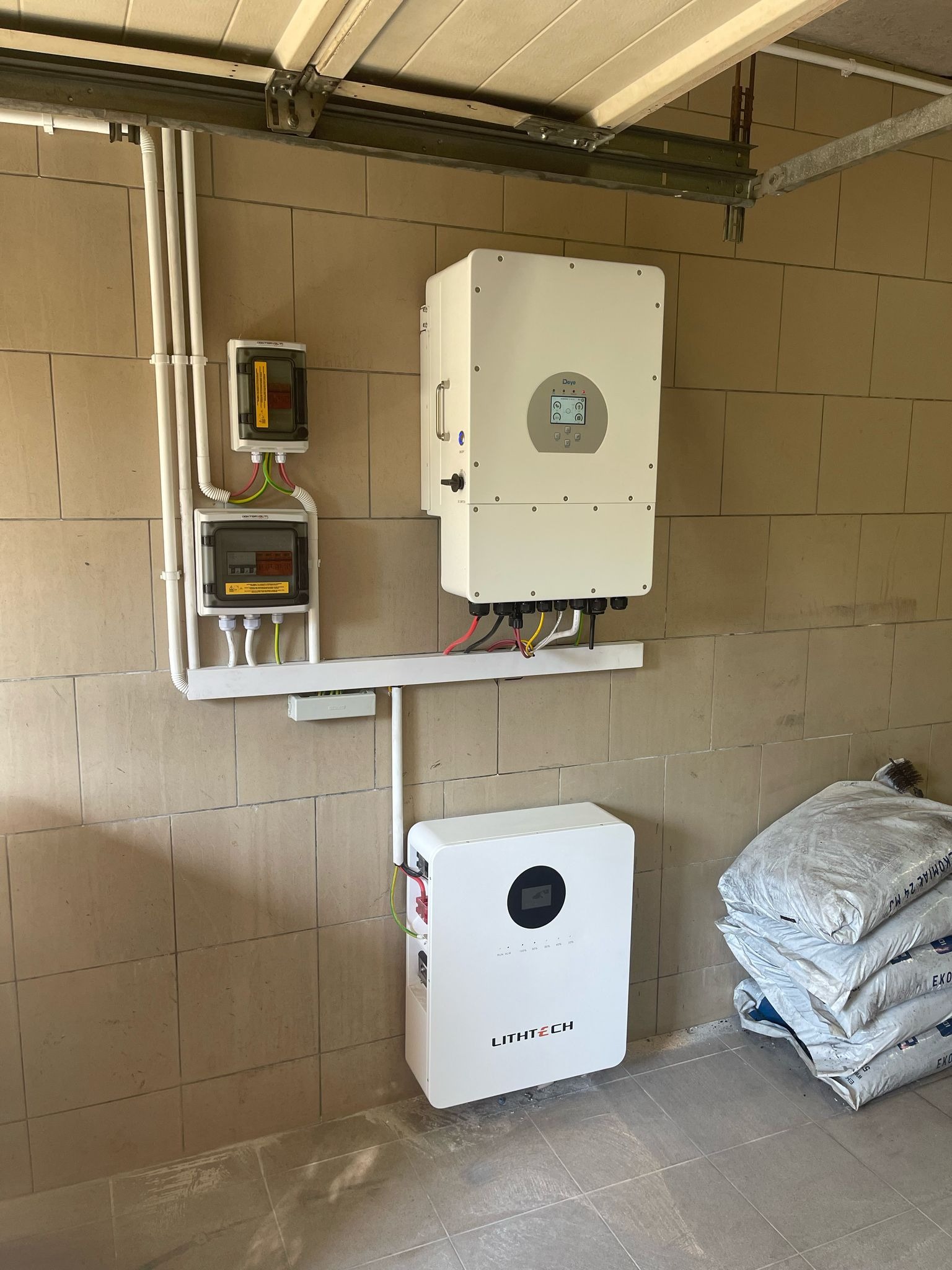 Kompletna instalacja z białym inwerterem i magazynem energii LITH TECH na beżowej ścianie z bloków. Widoczne kable, rury i skrzynki elektryczne. Podłoga wyłożona płytkami.