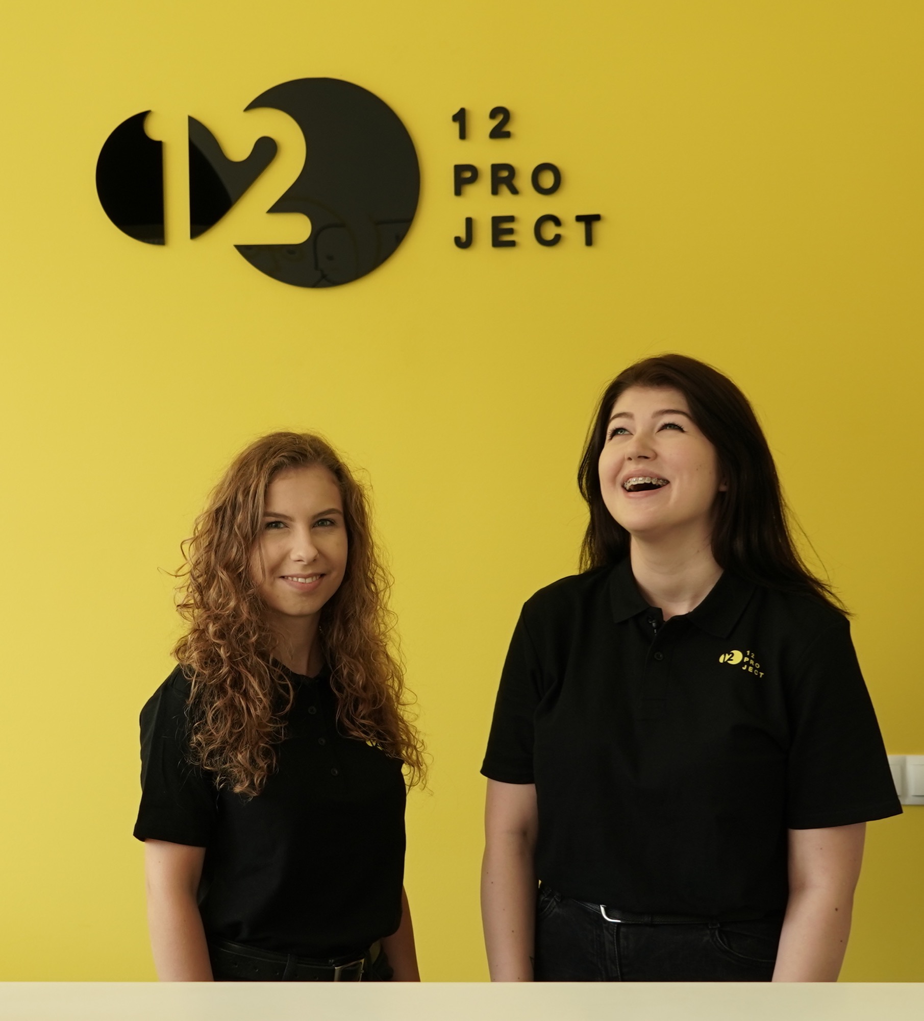 Dwie uśmiechnięte kobiety w czarnych koszulkach polo z logo firmy na tle żółtej ściany z napisem '12 PROJECT'.