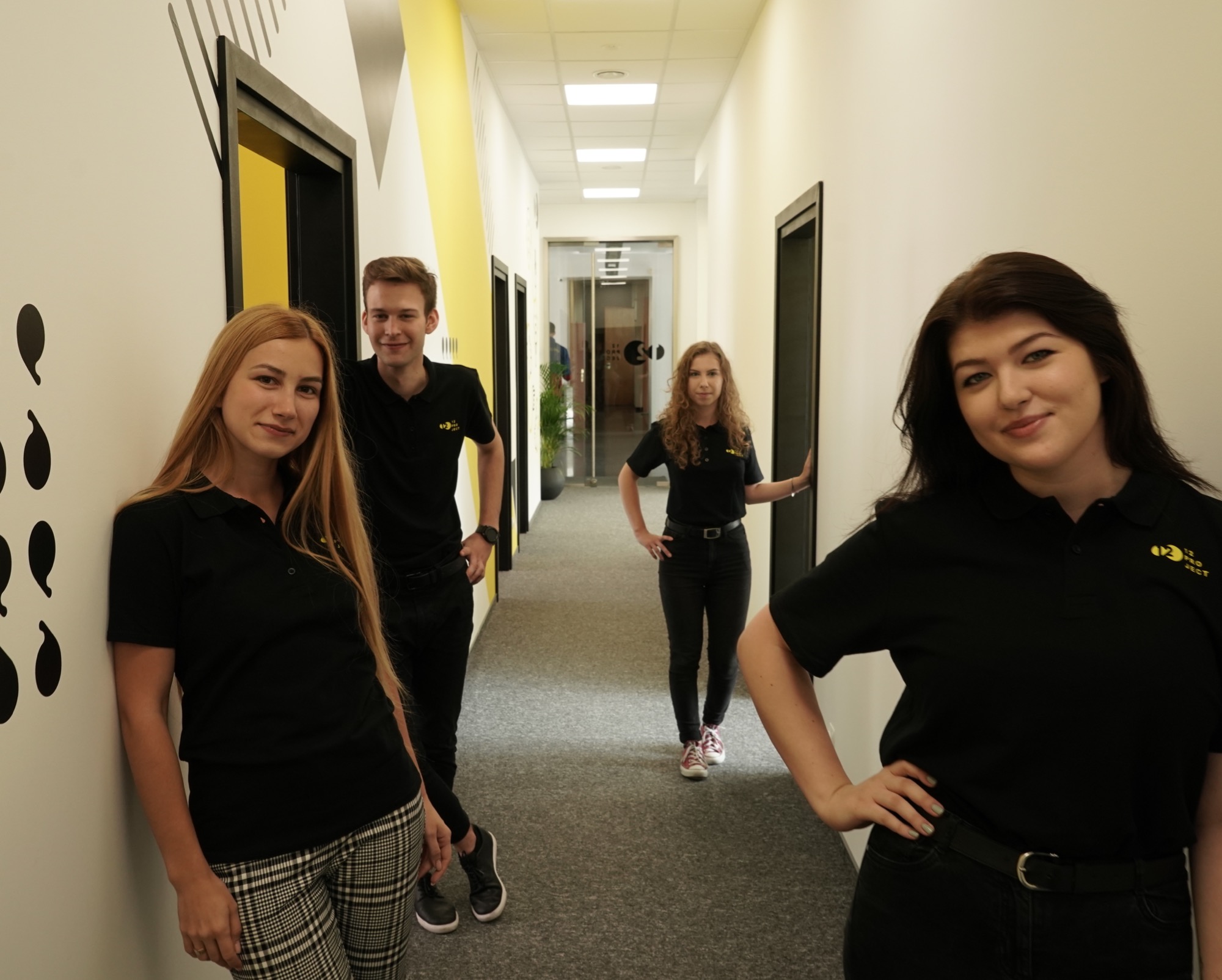 Zespół lektorów w Warszawie pozuje w korytarzu biura z czarno-żółtym wystrojem, ubrani w czarne koszulki polo z logo szkoły językowej.