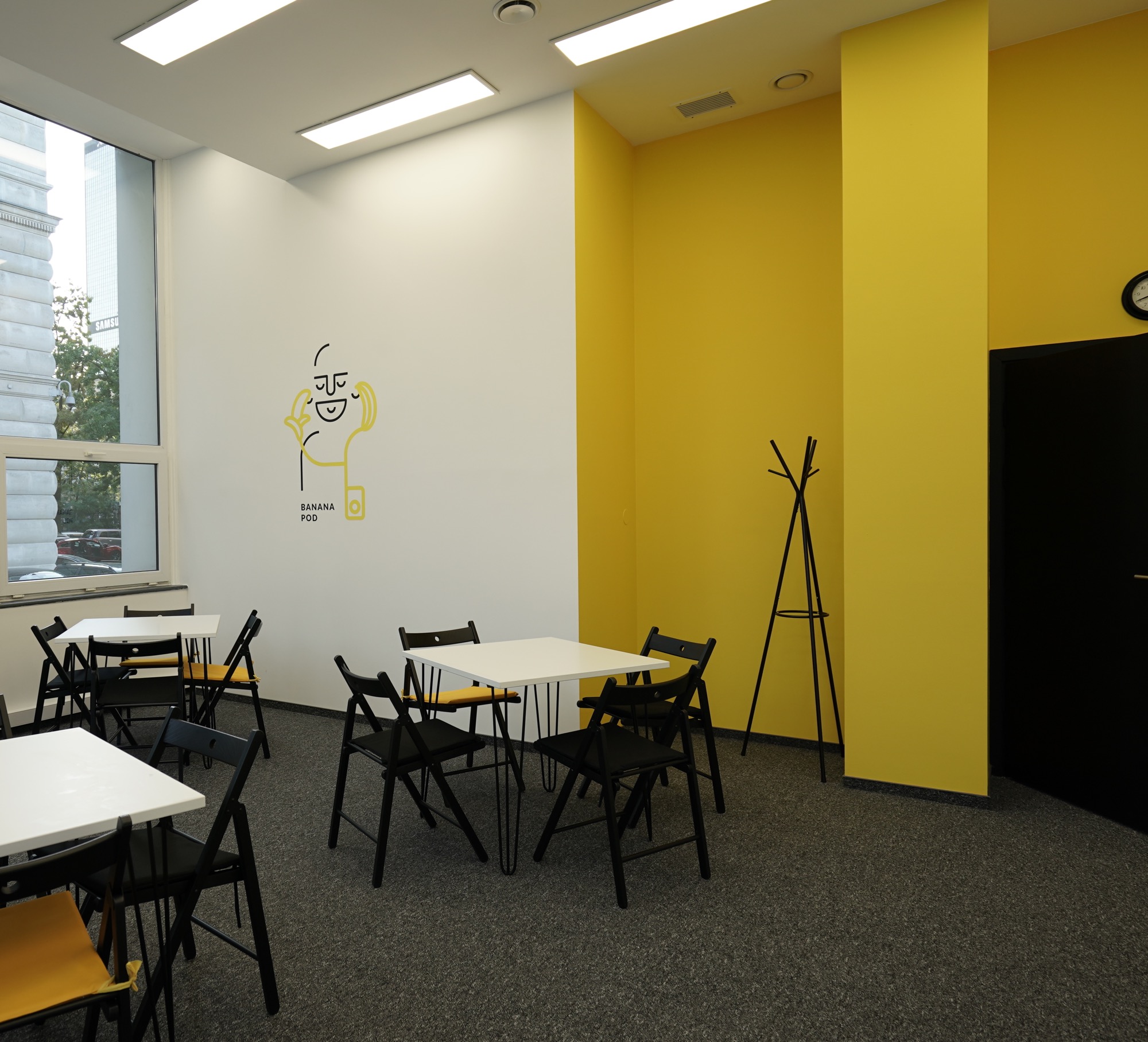 Jasne wnętrze z białymi stolikami, czarnymi krzesłami z żółtymi akcentami, żółtą ścianą i minimalistycznym rysunkiem na ścianie, prawdopodobnie sala lekcyjna lub przestrzeń coworkingowa.