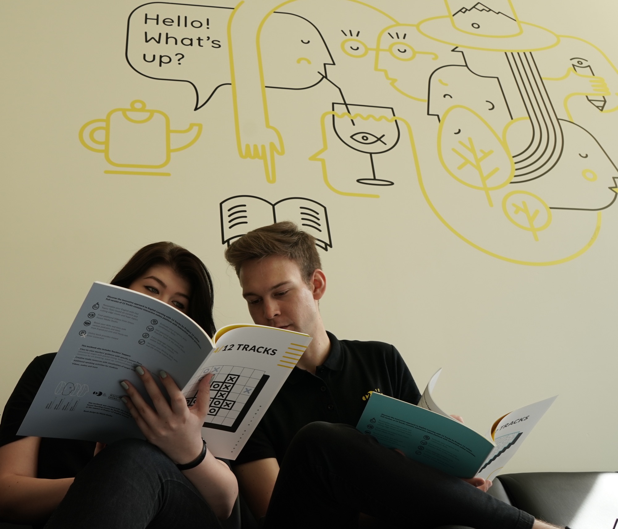 Dwoje młodych ludzi czyta podręczniki '12 Tracks' w nowoczesnym wnętrzu z ilustracjami na ścianie, prezentujące innowacyjne podejście do nauki języka angielskiego.