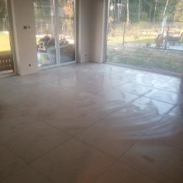 REM-BUD PIOTR SIKORA - Jasne, świeżo położone płytki podłogowe w przestronnym salonie z dużymi oknami wychodzącymi na ogród. Widoczne niedokończone ściany, przygotowane do malowania.