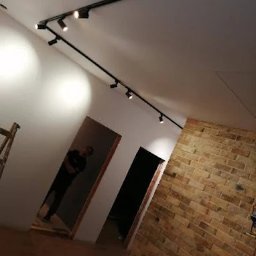 REM-BUD PIOTR SIKORA - Nowoczesne oświetlenie szynowe z reflektorami w świeżo wykończonym wnętrzu z ceglaną ścianą i drewnianą podłogą. Widoczna osoba w tle.